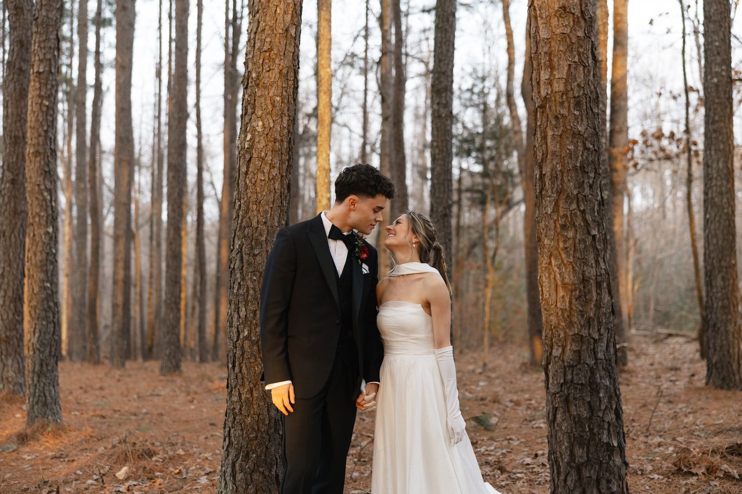 Newlyweds share a tender forehead kiss leaning against a pine tree in a sun-drenched autumn forest.