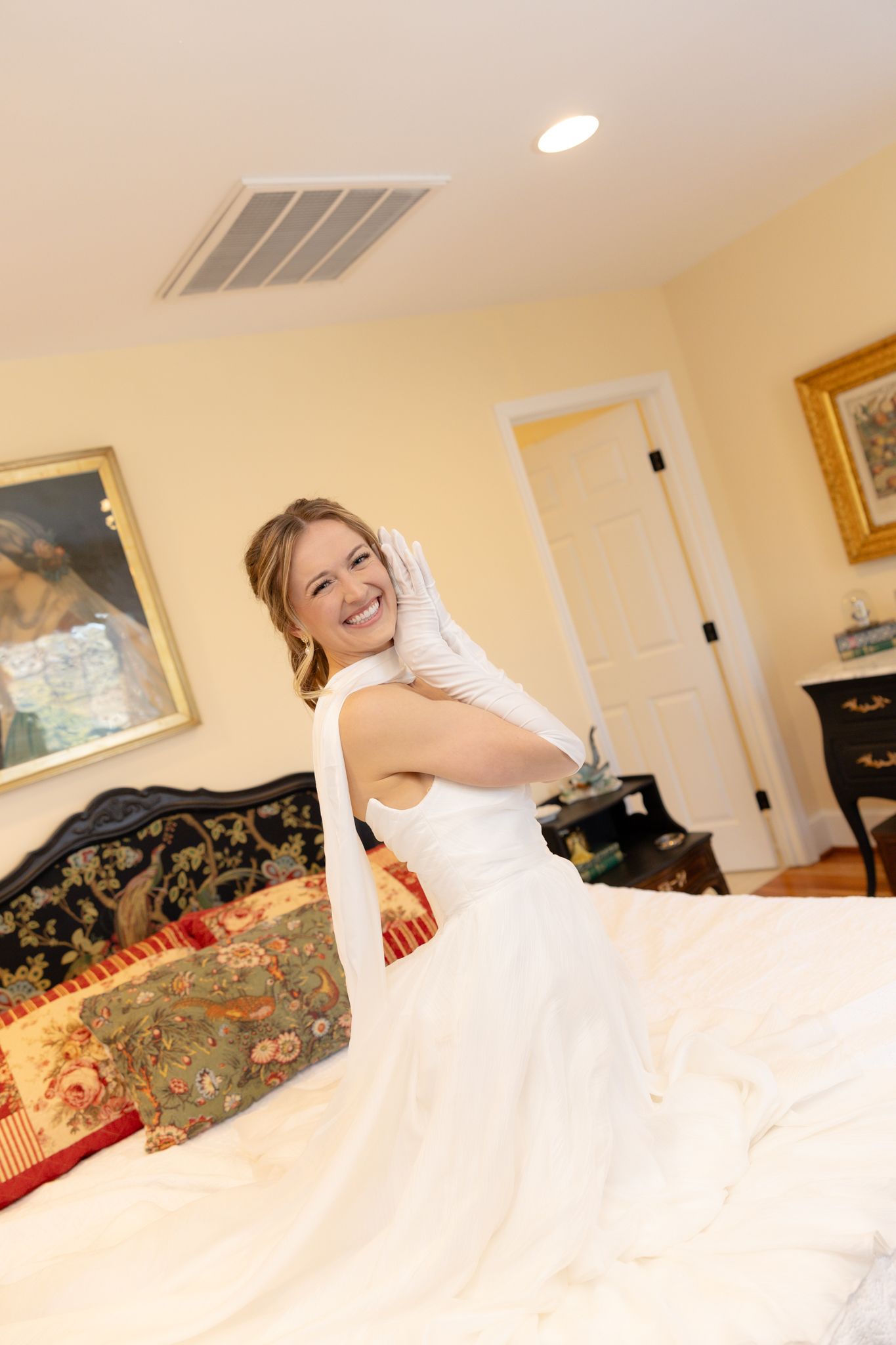 Joyful bride in white wedding dress and gloves smiling in a warmly decorated bedroom.