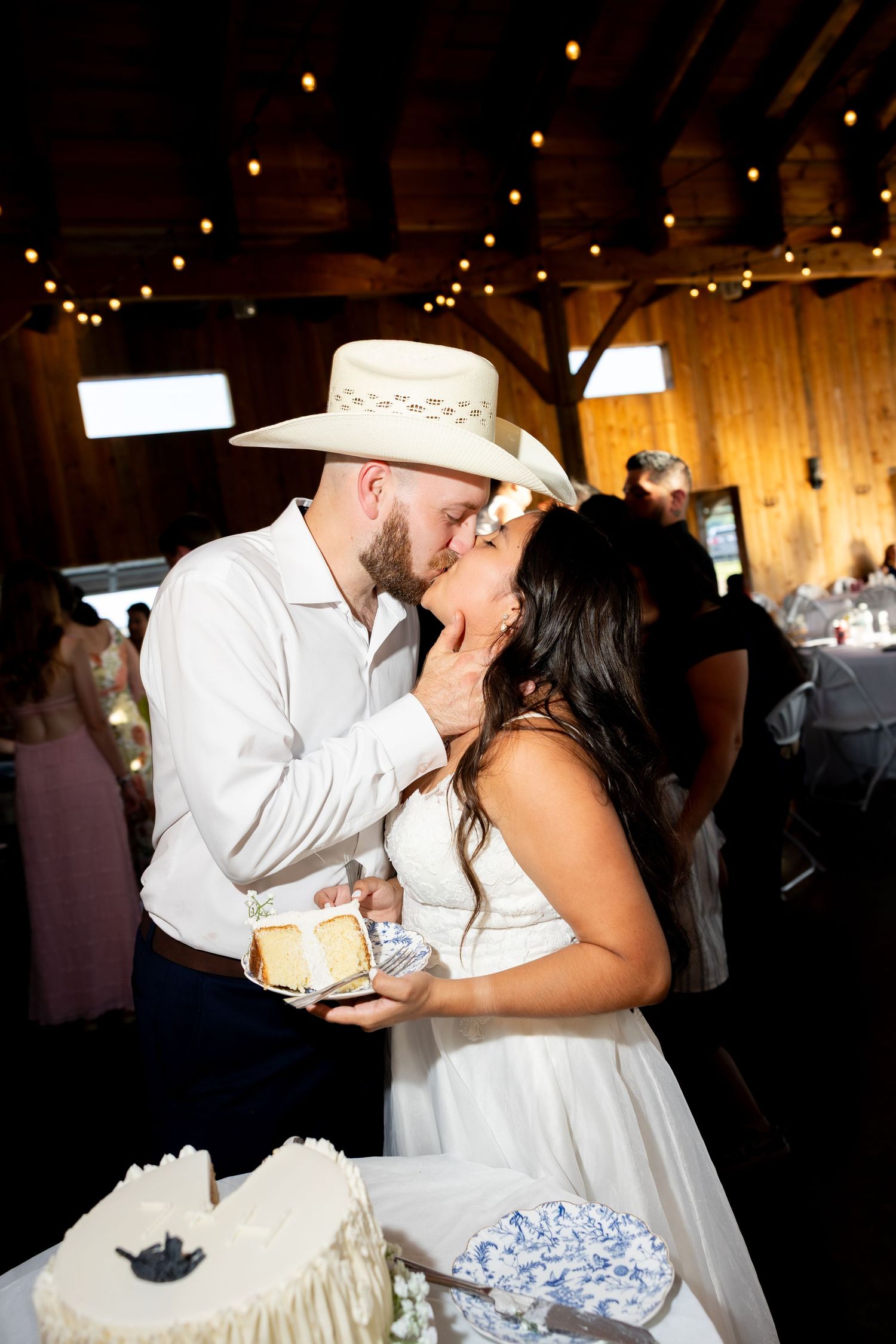 Bride and groom sharing a kiss while cutting their wedding cake in a rustic barn venue with string lights.