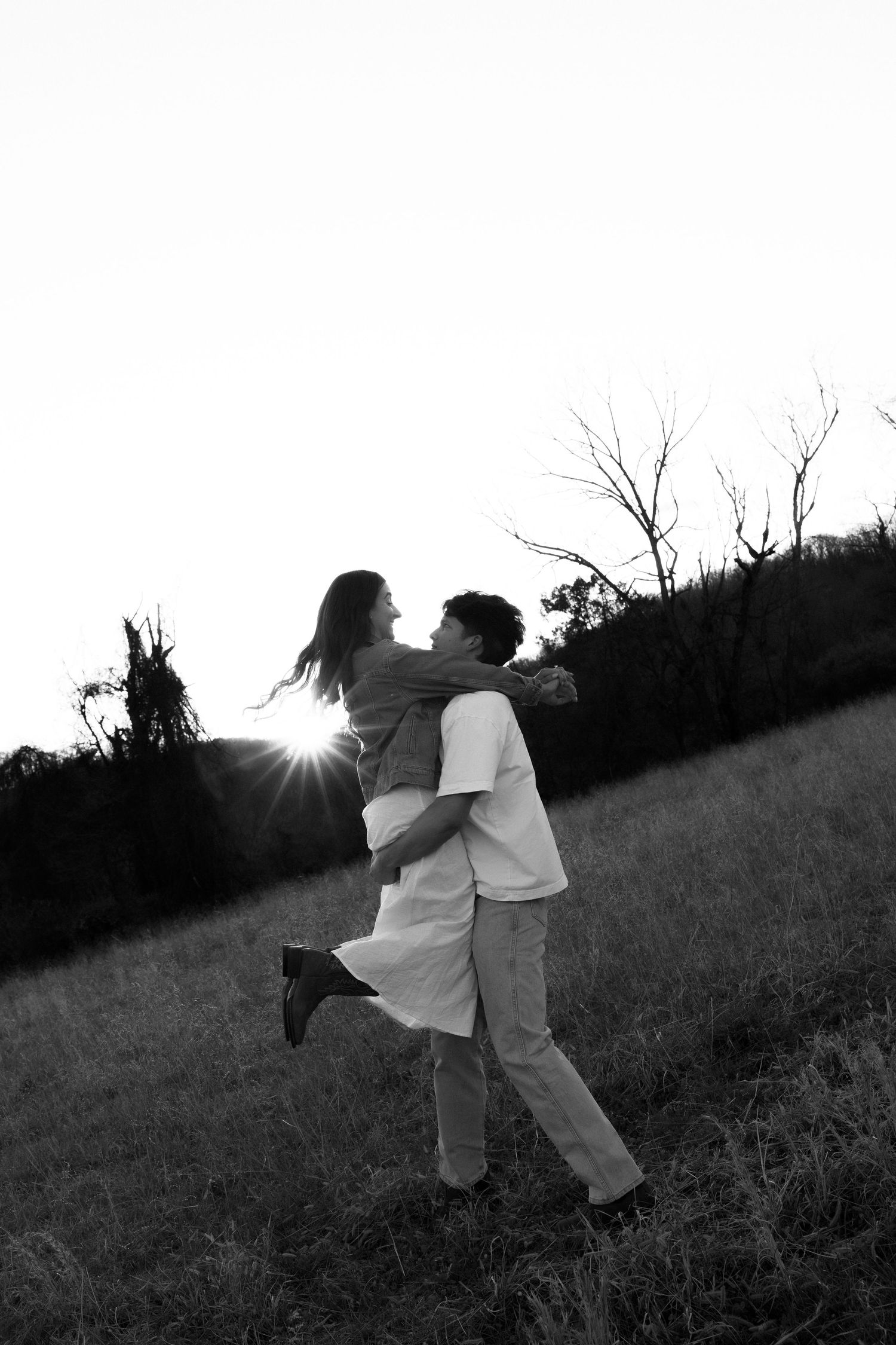 Black and white silhouette of a couple embracing in a sunlit field with a glowing sunset in the background.
