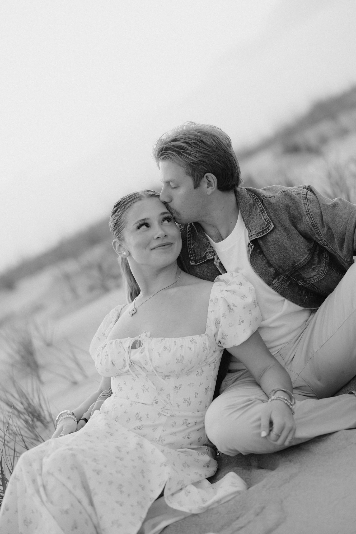 Black and white photo of a couple sitting on sand dunes, man kissing woman on cheek at sunset.