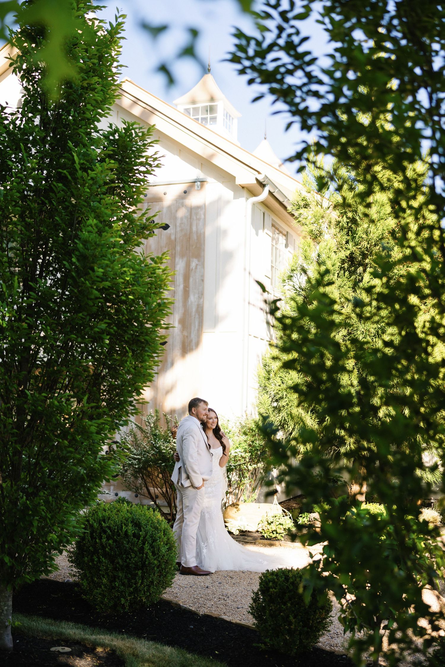 Bride and groom embracing in a lush garden framed by tall green hedges beside a white barn venue.