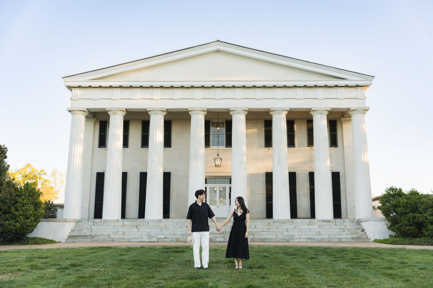 Couple holding hands in front of a grand white neoclassical building with tall columns at dusk.