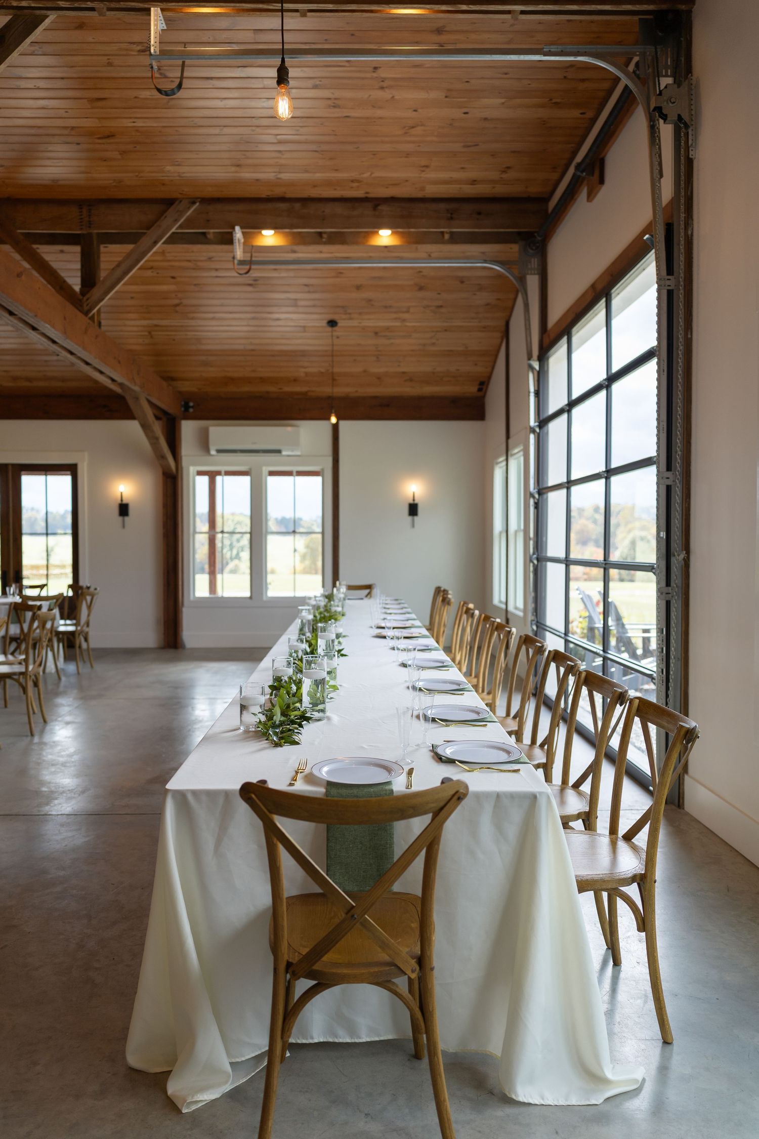 Elegant barn wedding venue with long white-draped table, wooden cross-back chairs, greenery centerpieces, and rustic wood-beam ceiling.