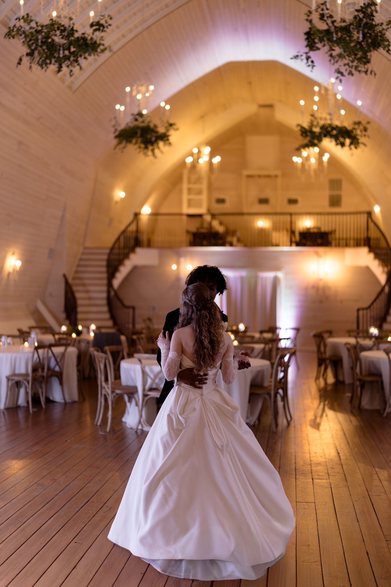 Bride and groom share their first dance in a warmly lit barn venue with vaulted ceilings.