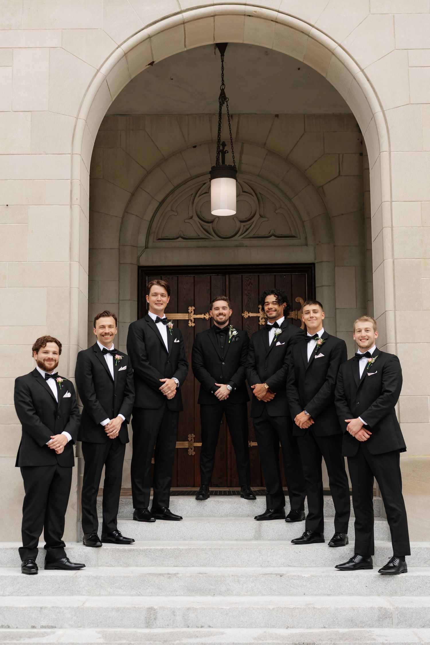 Seven groomsmen in black tuxedos pose on stone steps under an elegant arched doorway at a wedding venue.