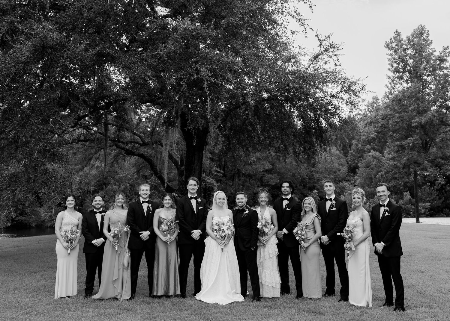 Black and white wedding party photo with bride, groom, bridesmaids, and groomsmen posing outdoors under large trees.