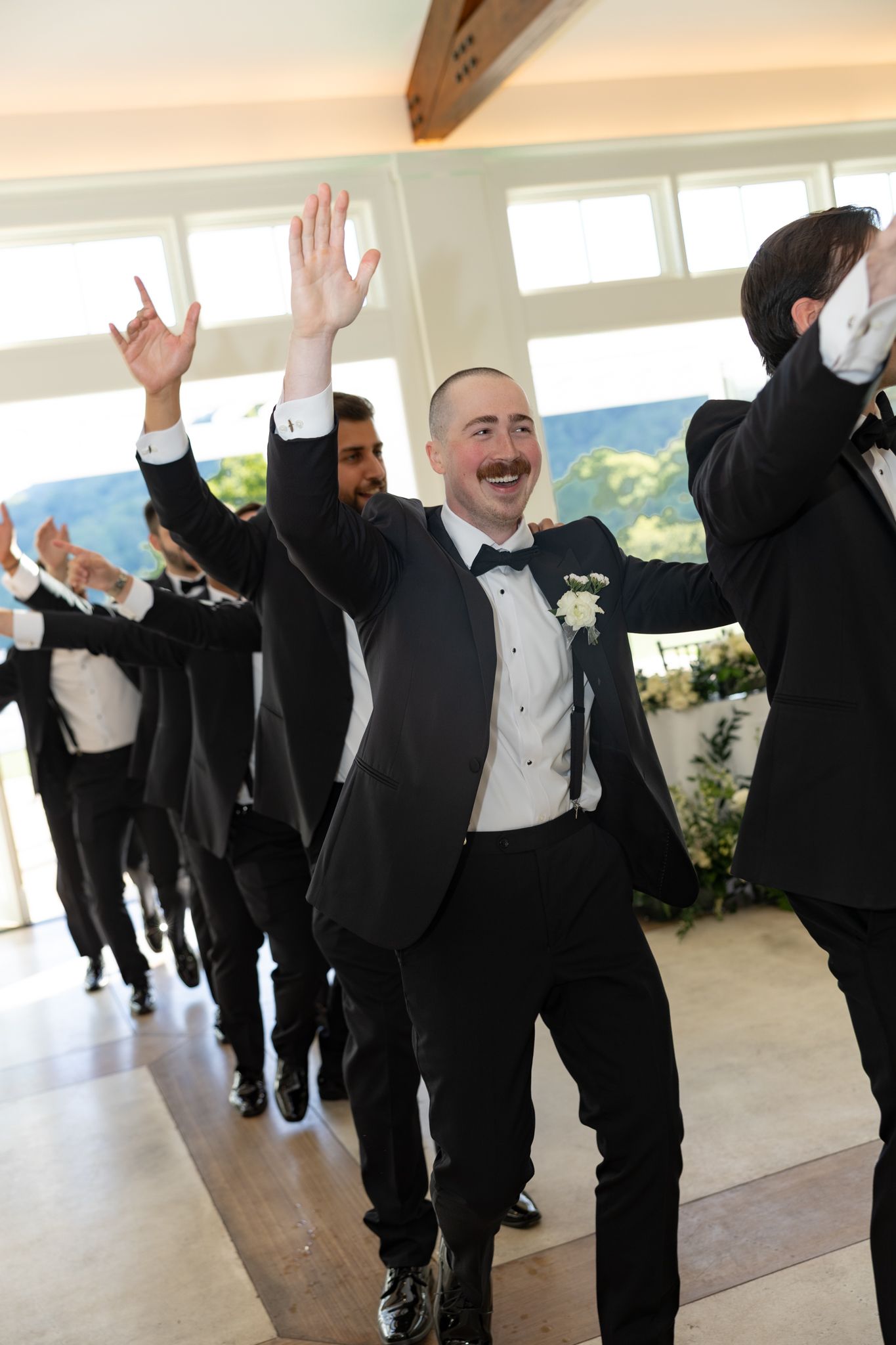 Groomsmen in black tuxedos celebrate with raised hands at a wedding reception in an elegant venue.