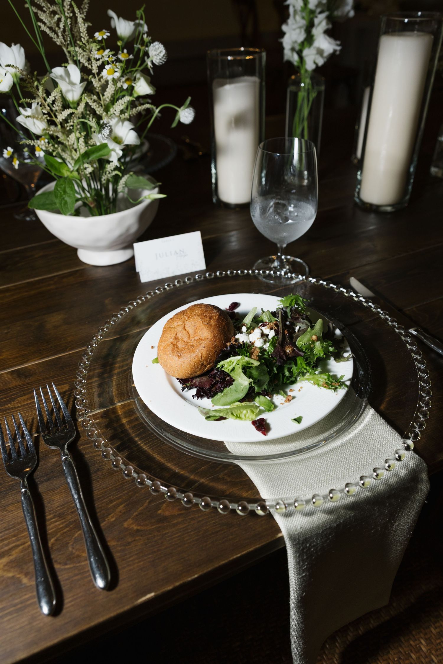 Elegant wedding reception place setting with salad, bread roll, glass charger plate, and white floral centerpiece.