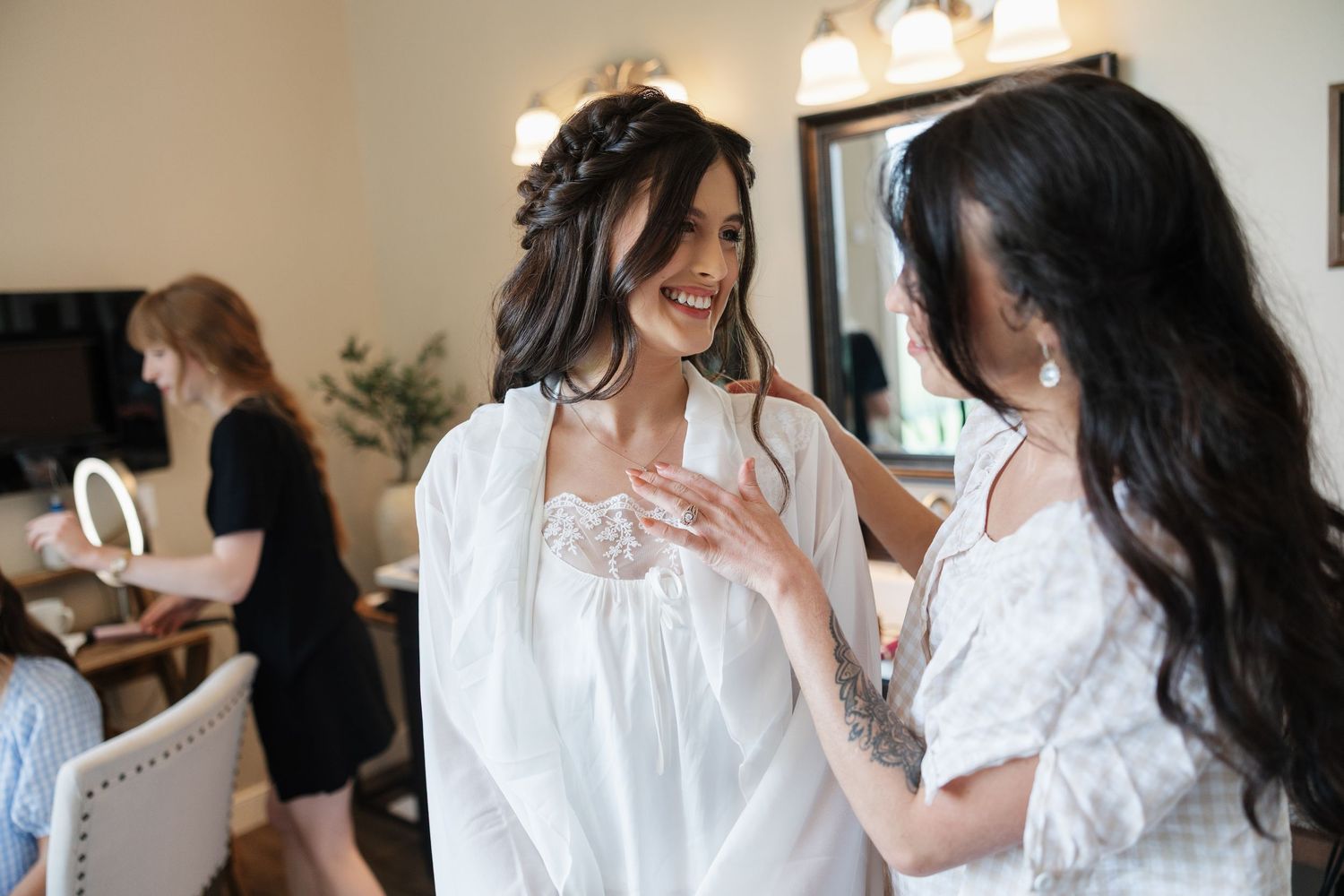 Bride in white robe smiling with bridesmaid during wedding preparations in a bridal suite.