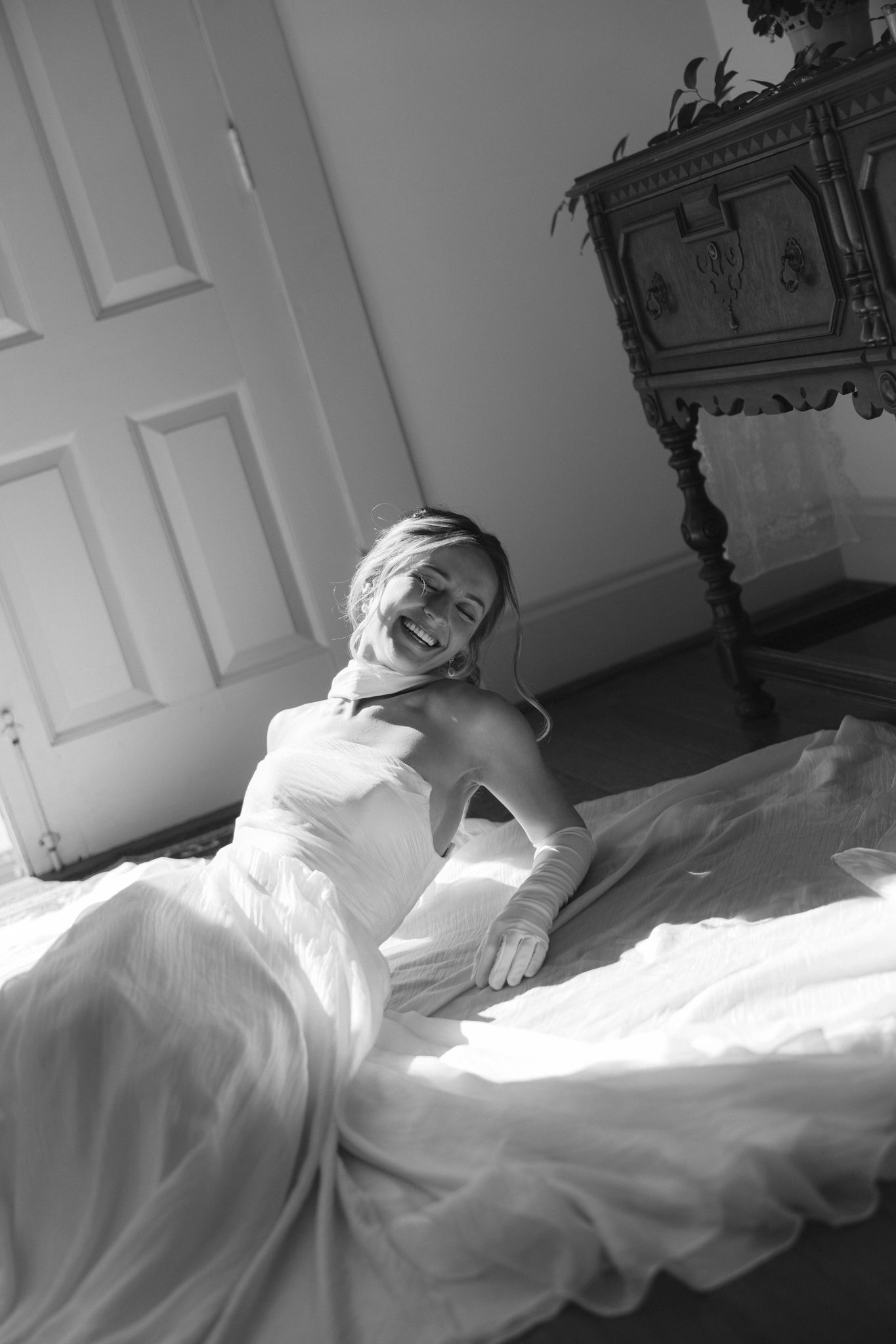 Black and white photo of laughing bride in wedding gown sitting on sunlit floor.