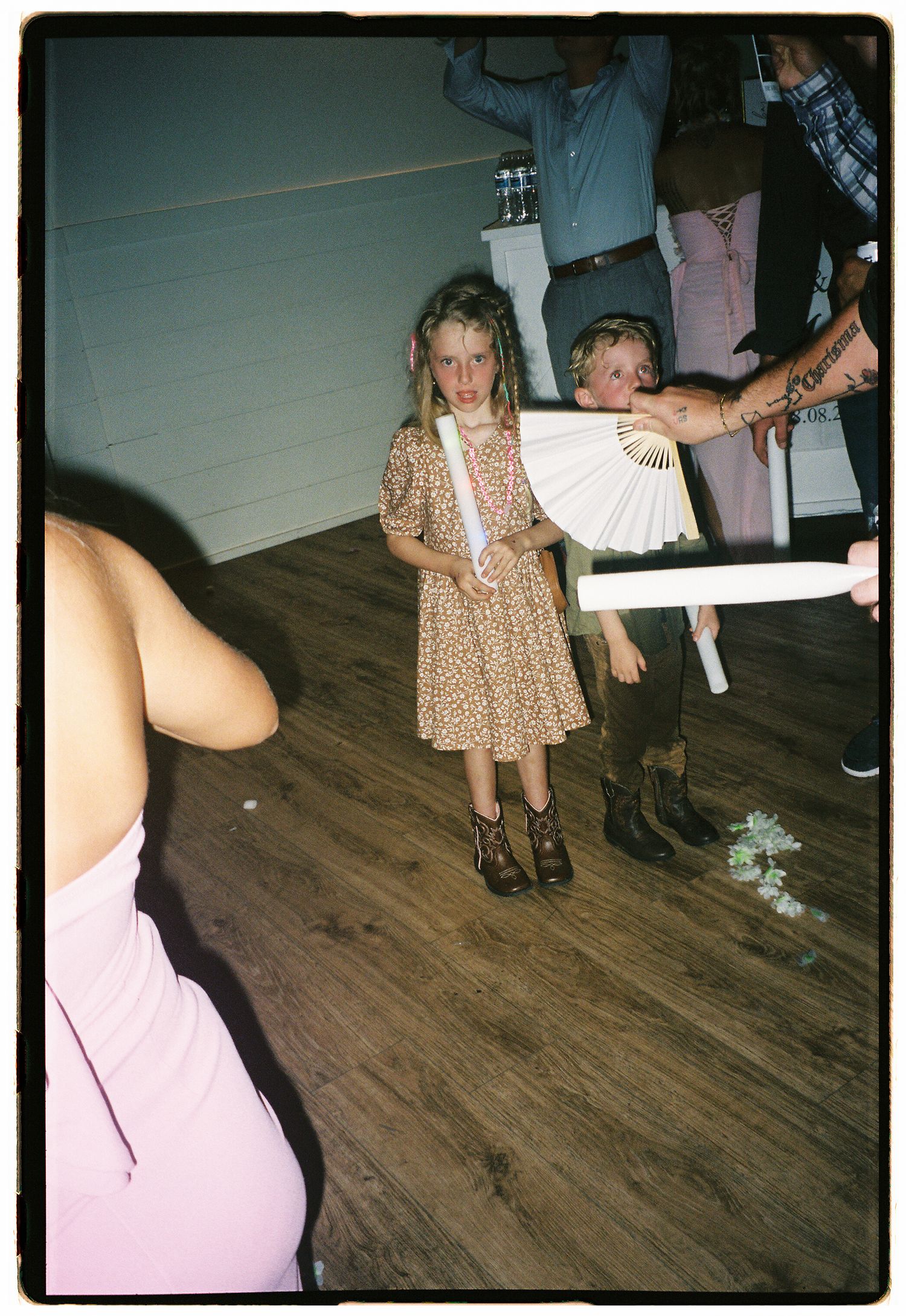 Two young children in cowboy boots hold white fans at a rustic indoor party with wood floors.