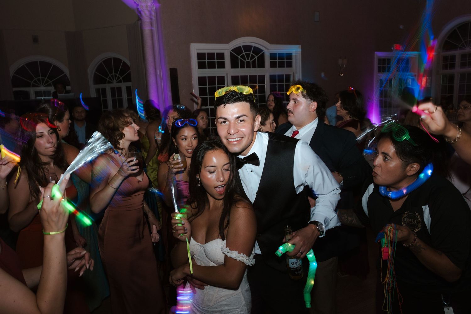 Bride and groom pose with guests holding glow sticks at a lively wedding reception dance party.