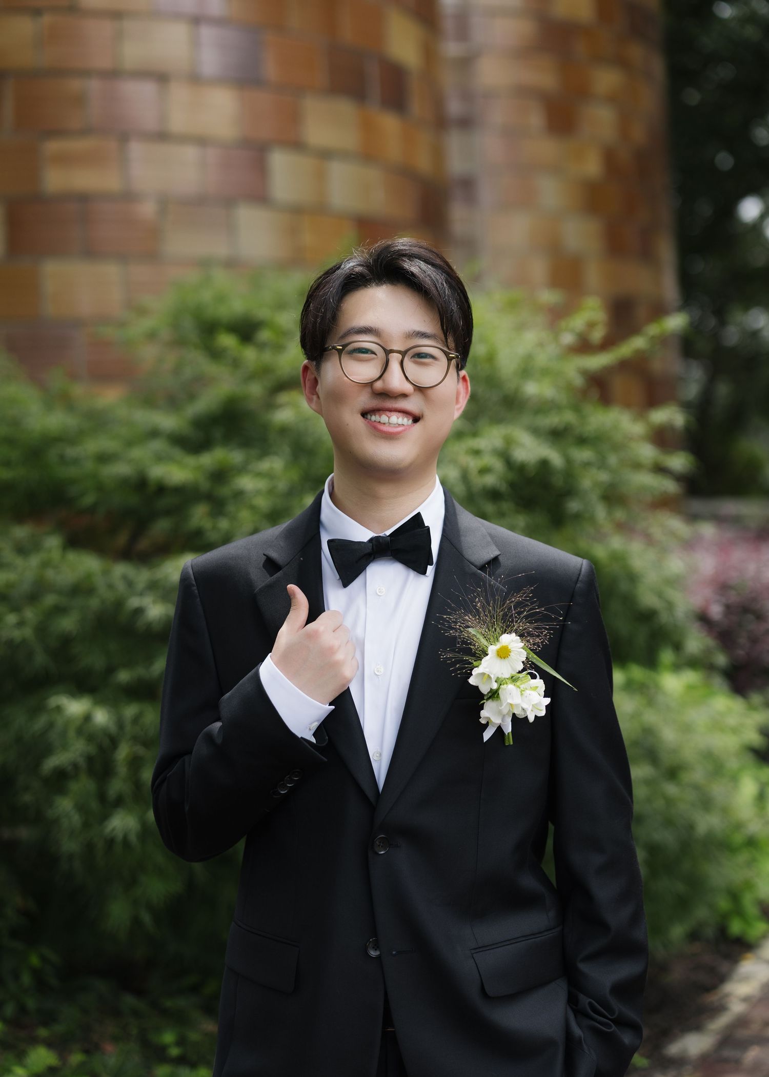Young man in black tuxedo with bow tie and white boutonniere giving thumbs up, smiling outdoors.