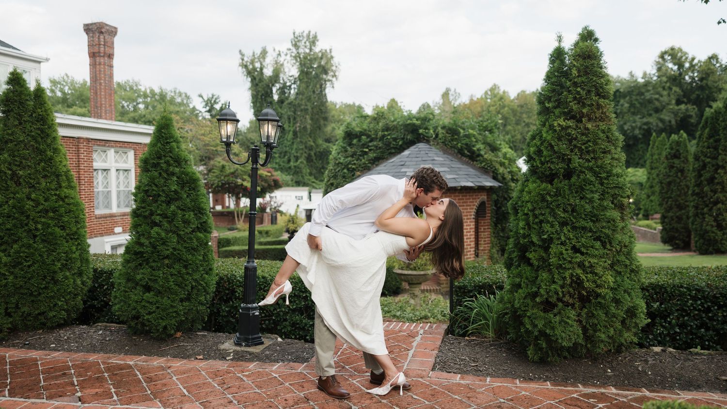 Bride and groom share a romantic dip kiss on a brick pathway surrounded by lush greenery at an elegant outdoor wedding venue.