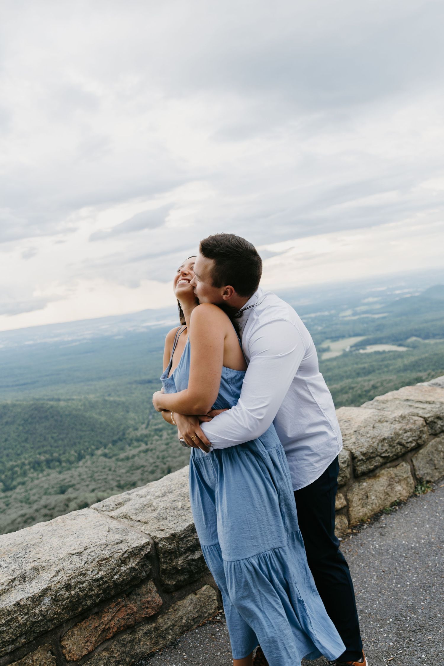 Man embracing woman from behind on mountain overlook under cloudy sky.