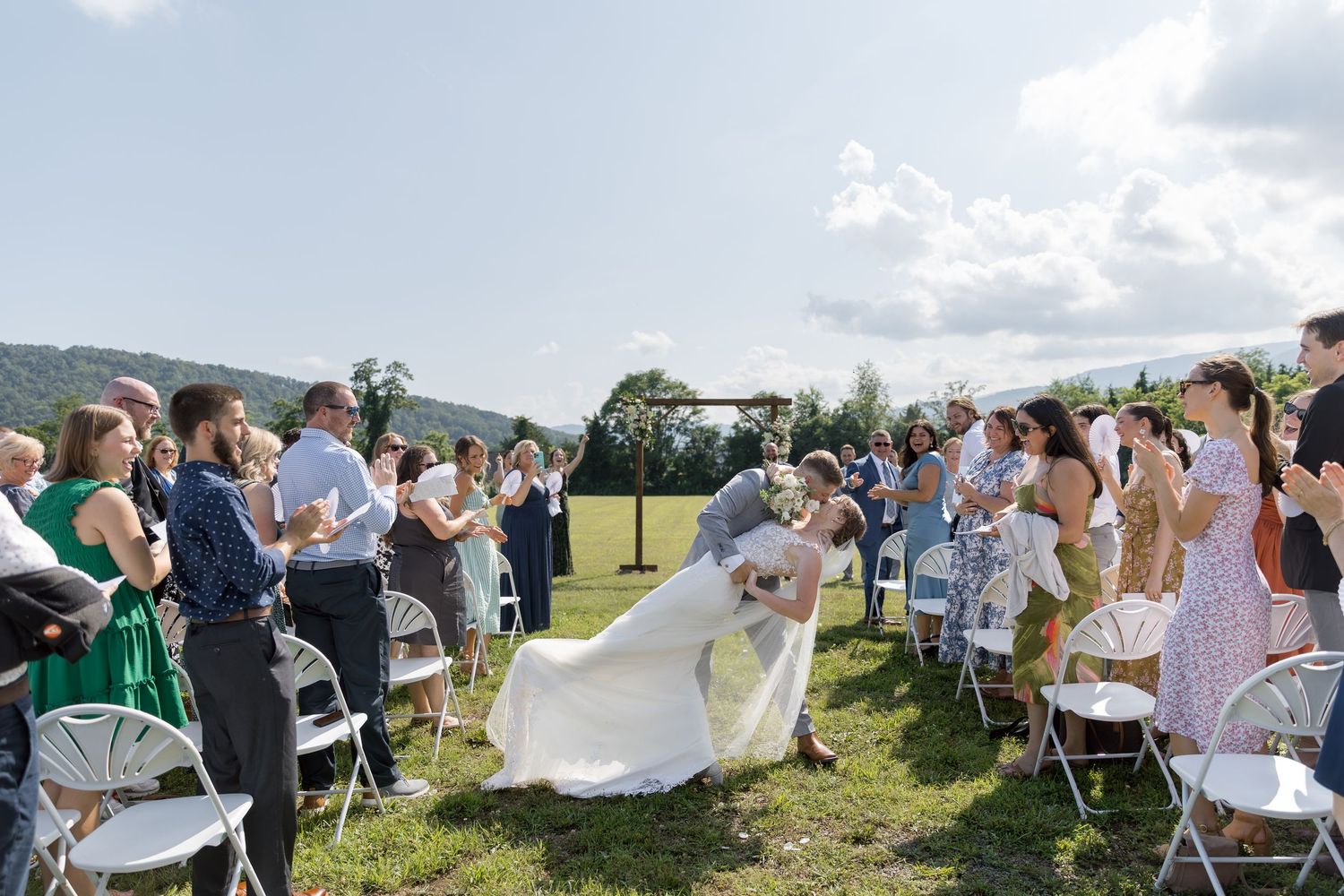 Bride and groom share a romantic dip kiss while wedding guests cheer along the aisle outdoors.