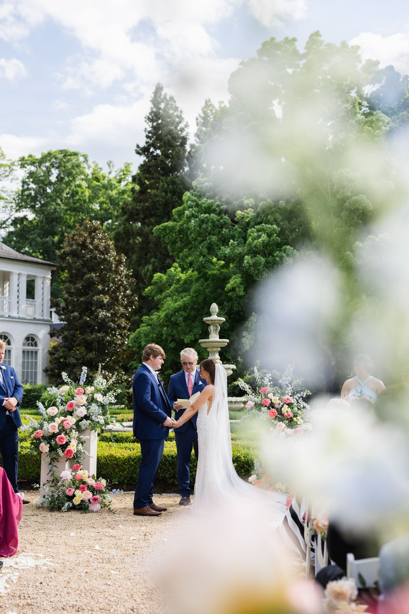 Outdoor wedding ceremony in elegant garden with floral arch, bride and groom exchanging vows beside a grand estate.