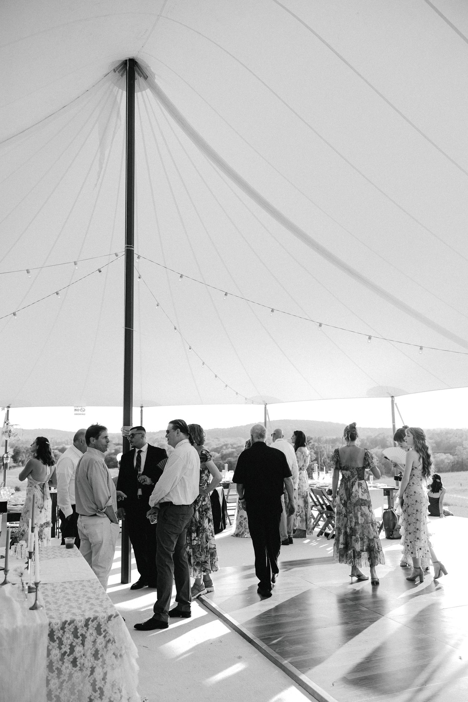 Black and white photo of wedding guests mingling under a large sailcloth tent with string lights.