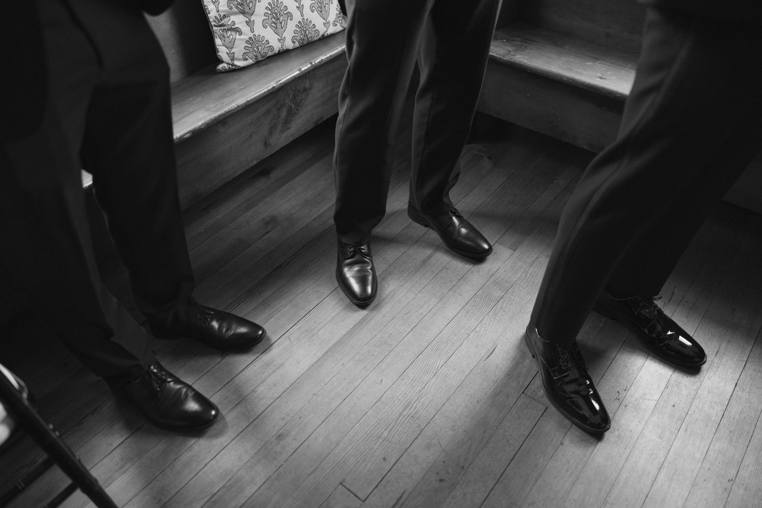 Black and white photo of groomsmen in dress shoes standing on hardwood floor.