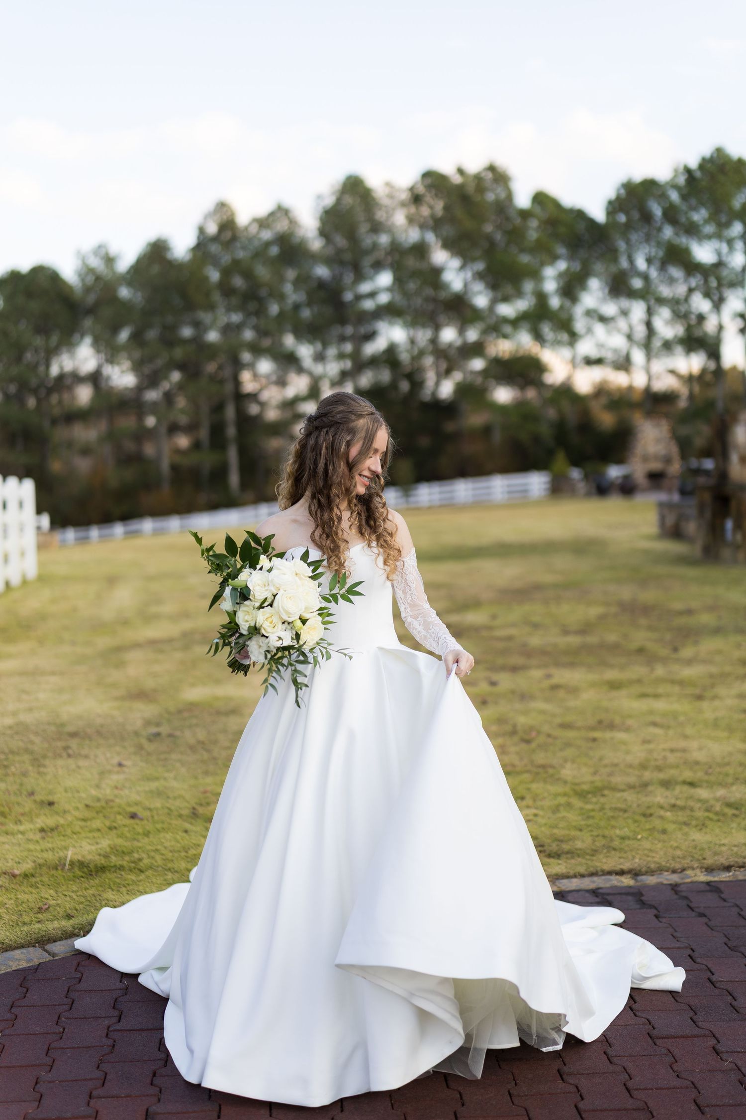 Bride in long-sleeve white ballgown holds white floral bouquet outdoors at scenic wedding venue.