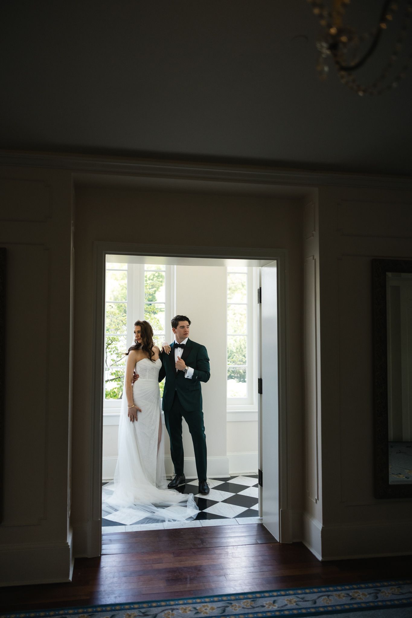 Bride in white gown and groom in black tuxedo stand in elegant doorway with checkered floor at wedding venue.