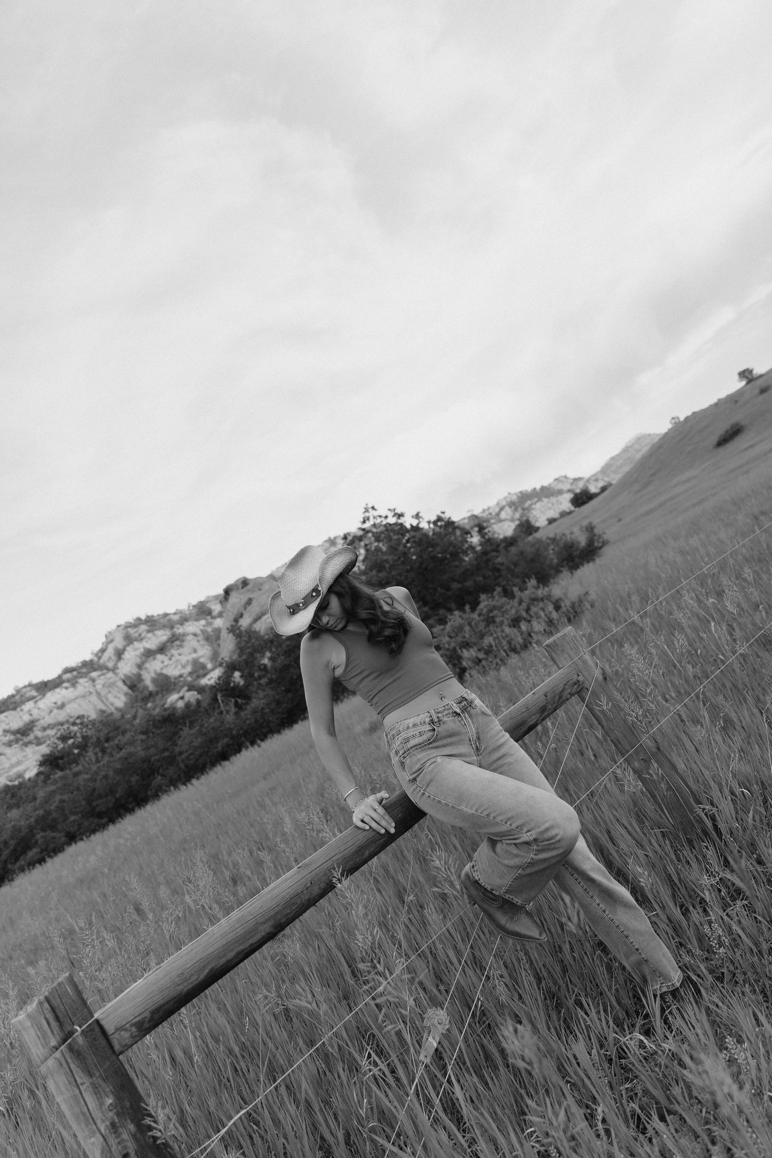 Black and white photo of woman in cowboy hat leaning on wooden fence in mountain meadow.