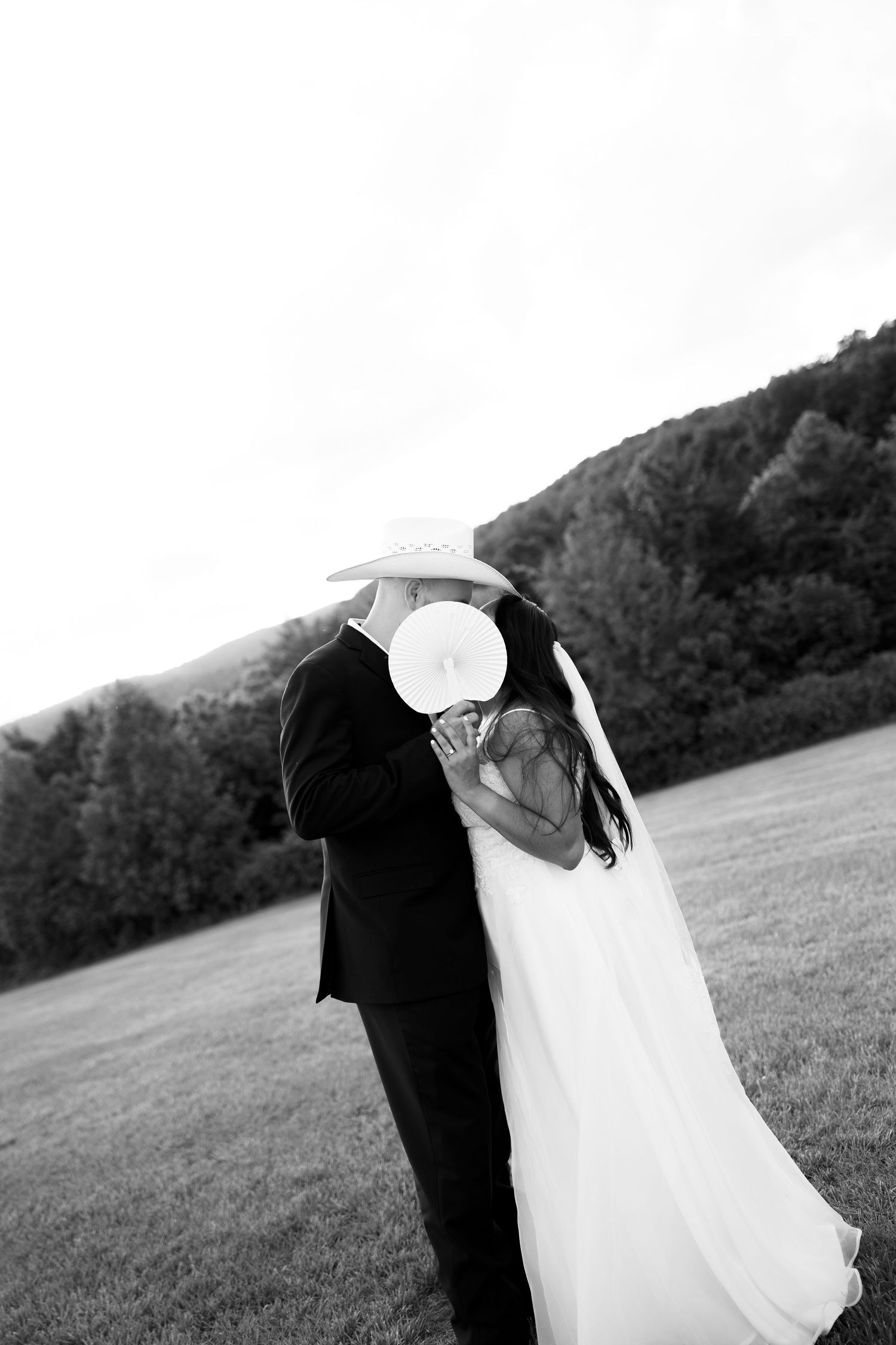 Black and white photo of a bride and groom sharing a kiss hidden behind a cowboy hat in an open field.