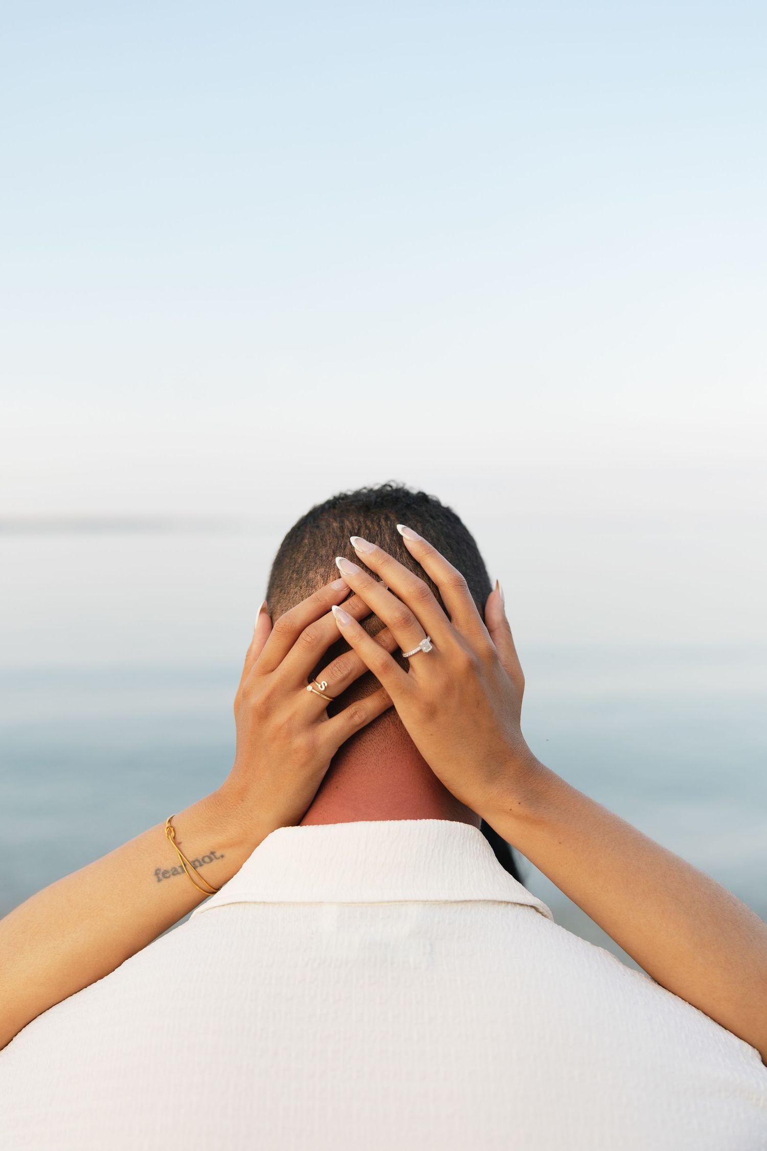 Woman's hands with engagement ring resting on man's head near calm ocean waters at dusk.