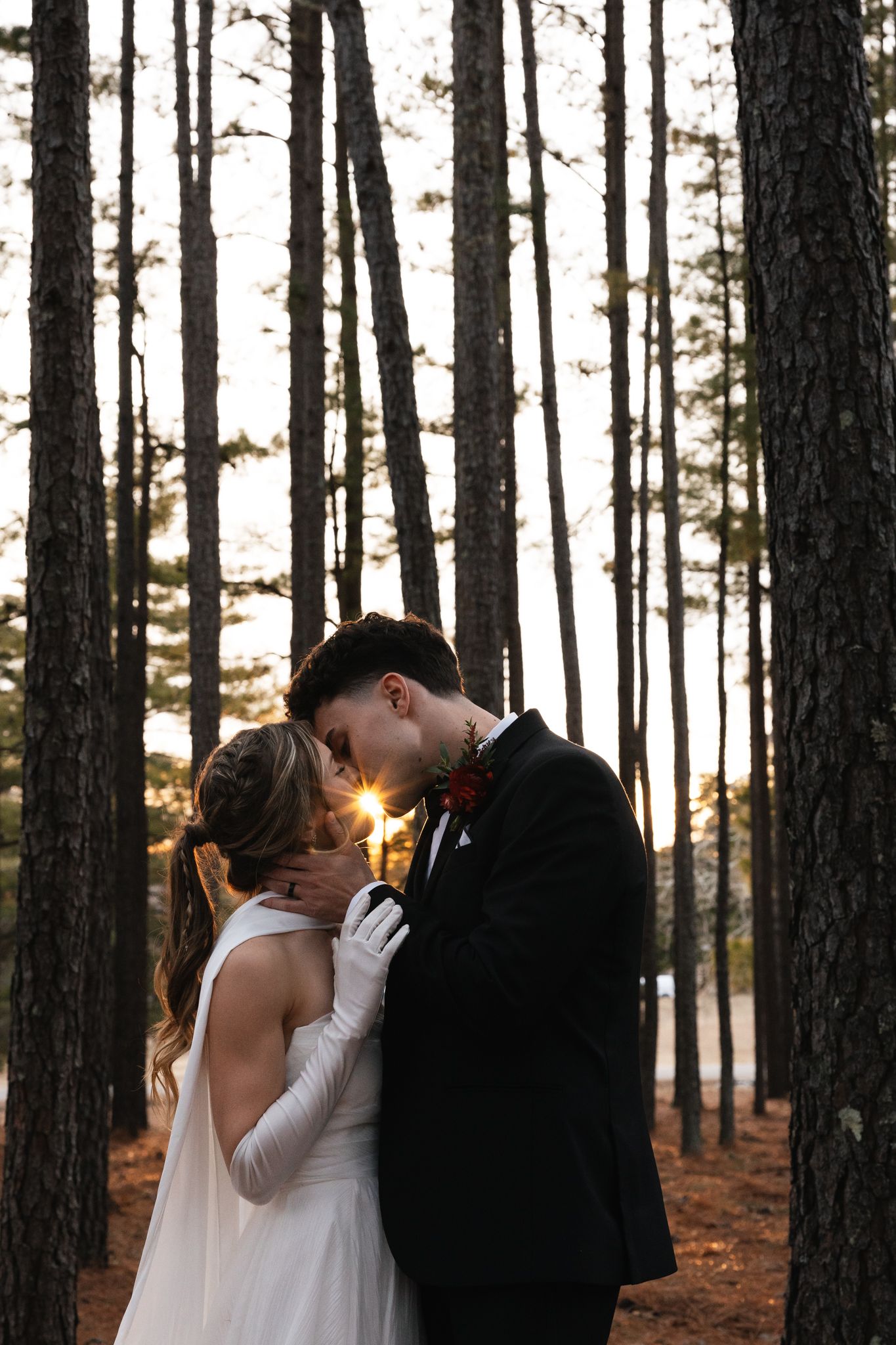 Bride and groom share romantic forehead touch in pine forest with golden sunset glowing behind them.