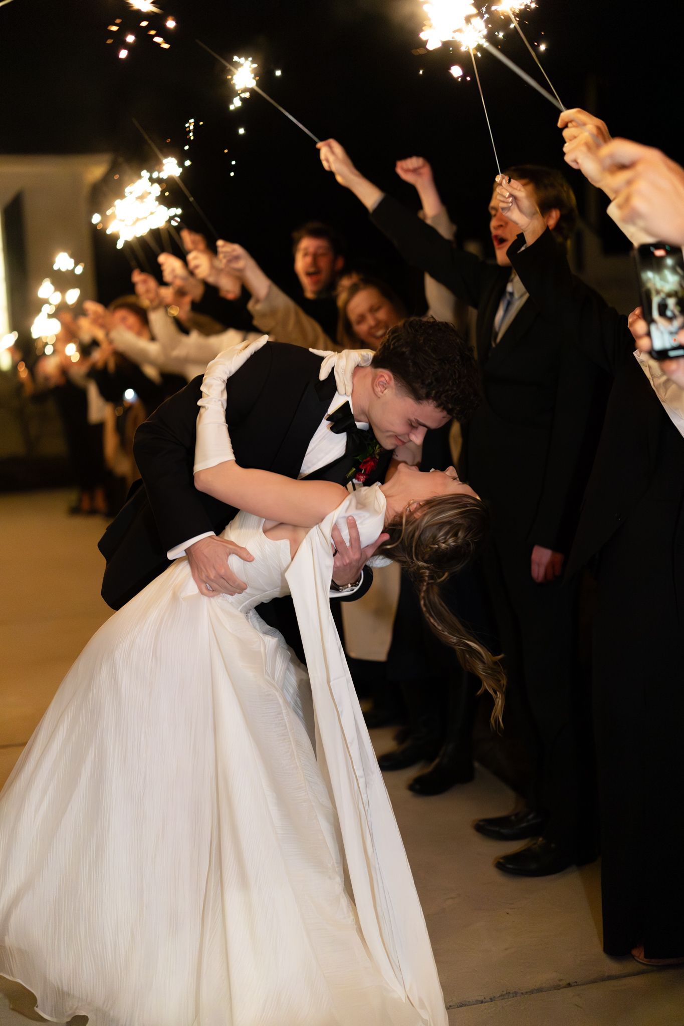 Groom dips bride for a passionate kiss during a sparkler exit as excited wedding guests celebrate around them.