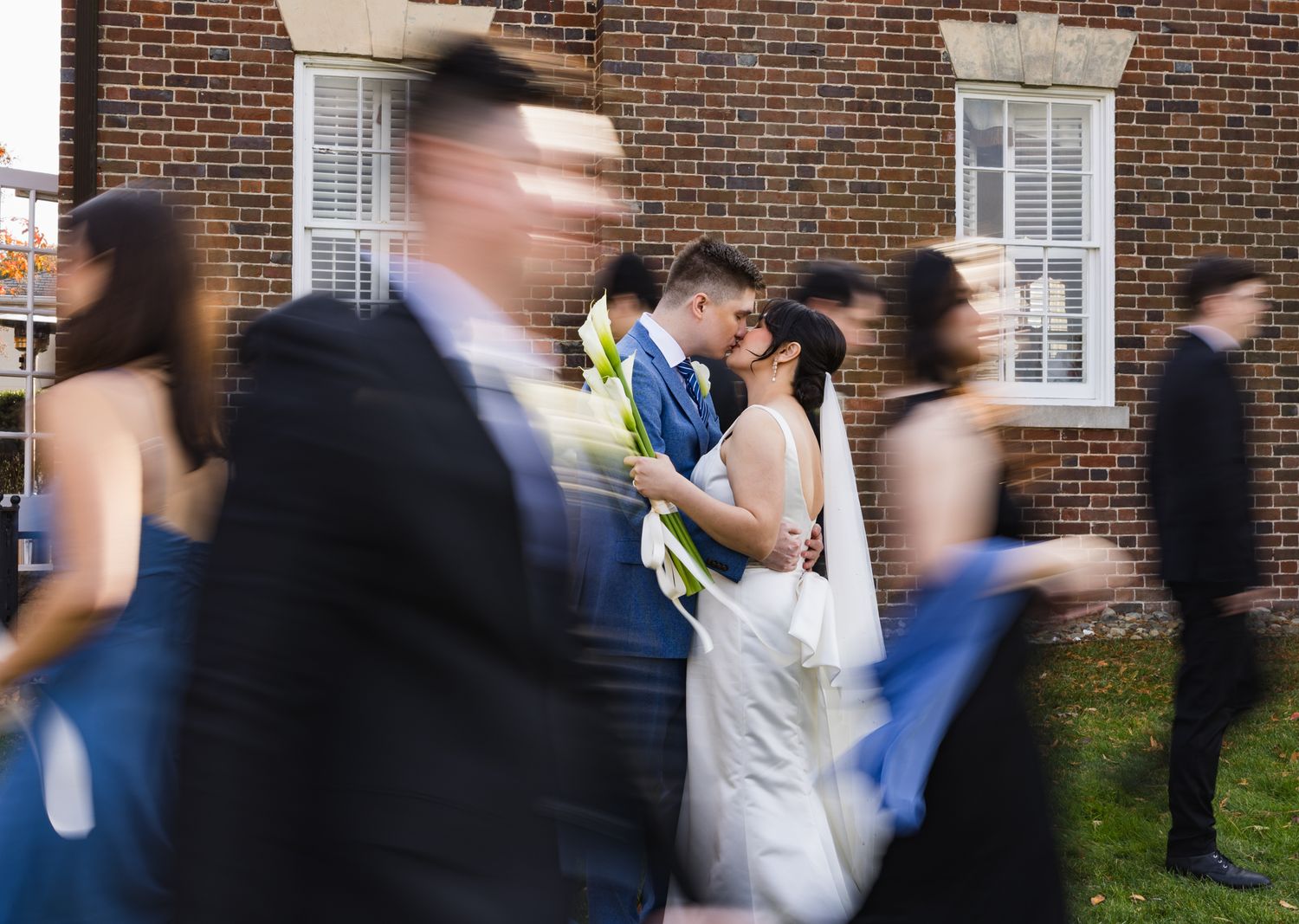 Bride and groom kiss as wedding guests walk past in motion blur outside brick building.