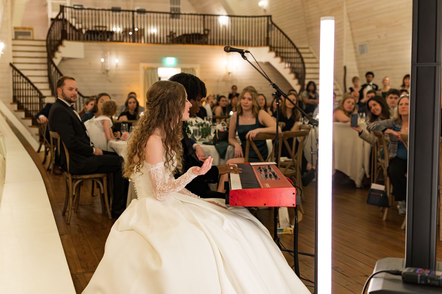 Bride in a ball gown plays a red piano for guests during a wedding reception performance.
