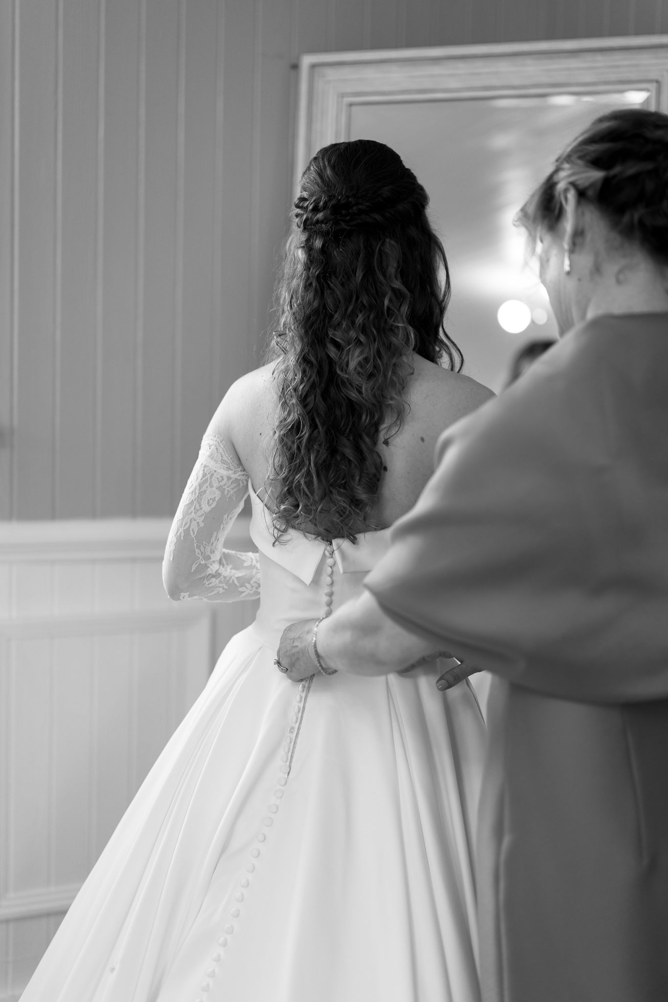 Black and white photo of bride in lace wedding gown being buttoned up with long curly hair.