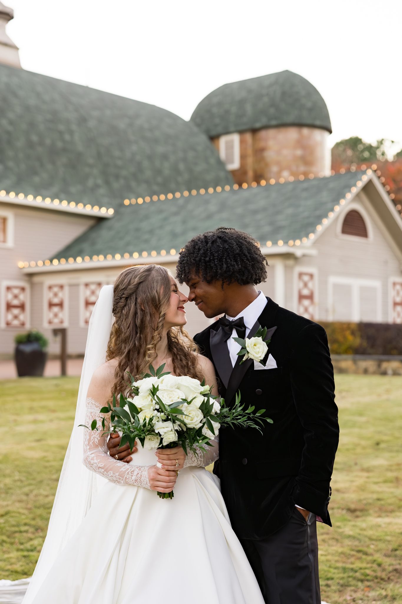 Interracial couple shares a romantic moment holding white roses outside a charming barn venue.