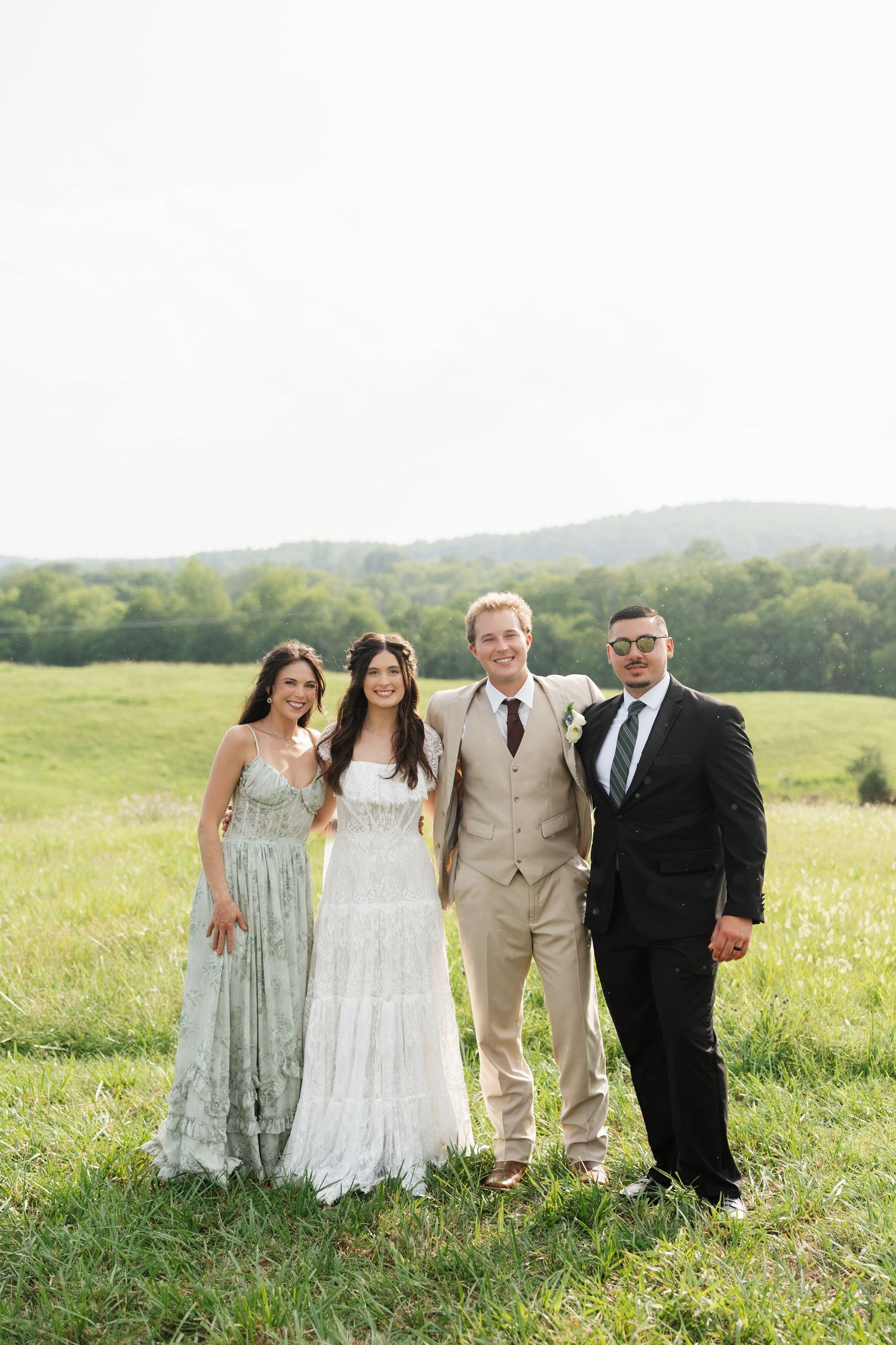 Wedding party of four posing in a green meadow, bride in white gown, groom in tan suit, outdoors.