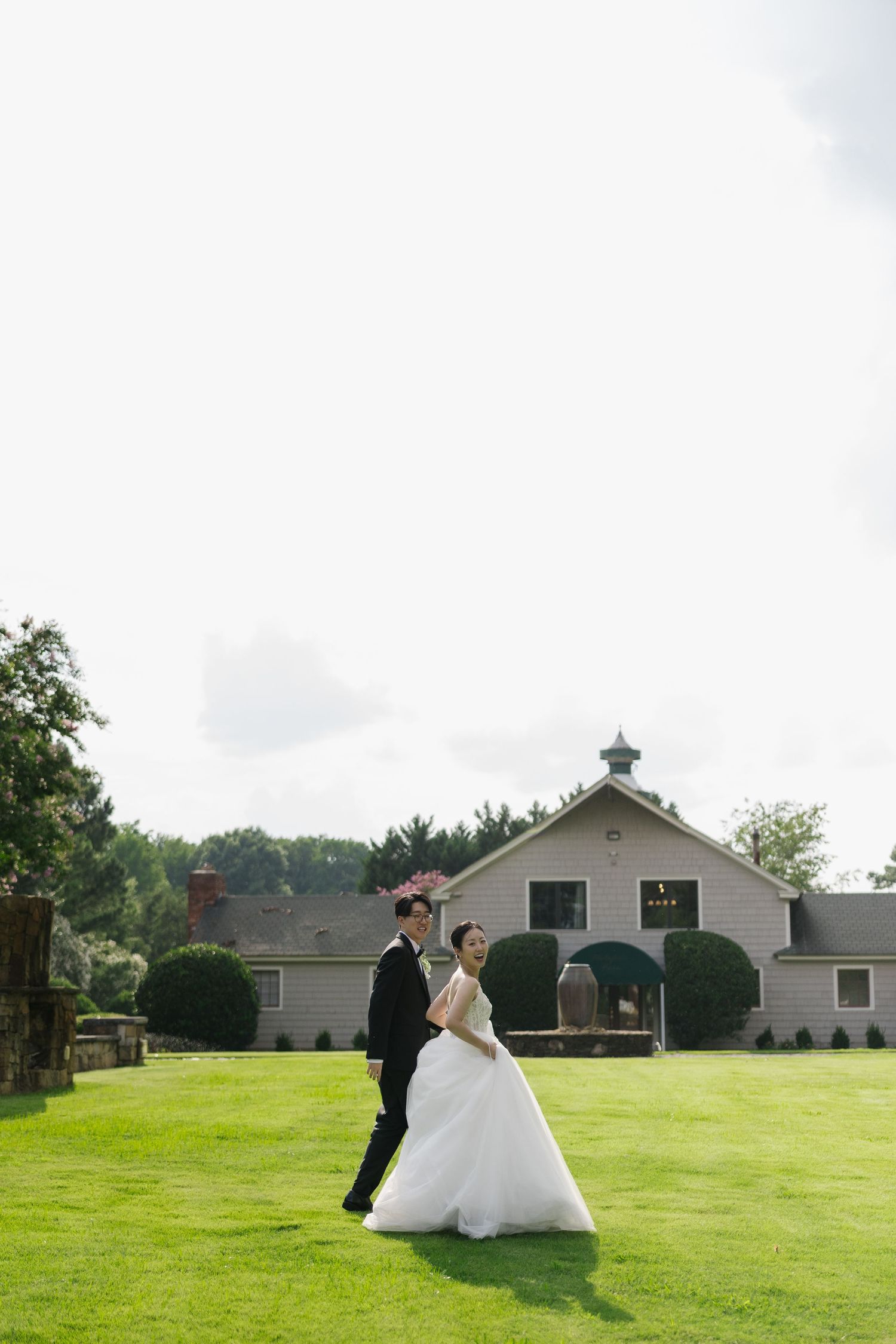 Bride and groom pose on lush green lawn before a charming white barn venue on their wedding day.