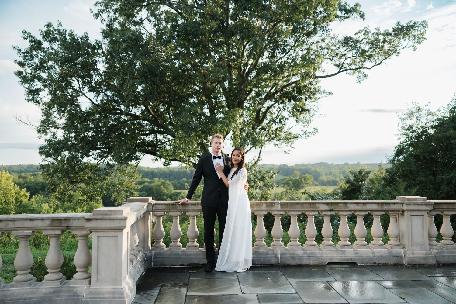 Bride and groom pose on stone terrace with elegant balustrade, lush green landscape and trees in background.