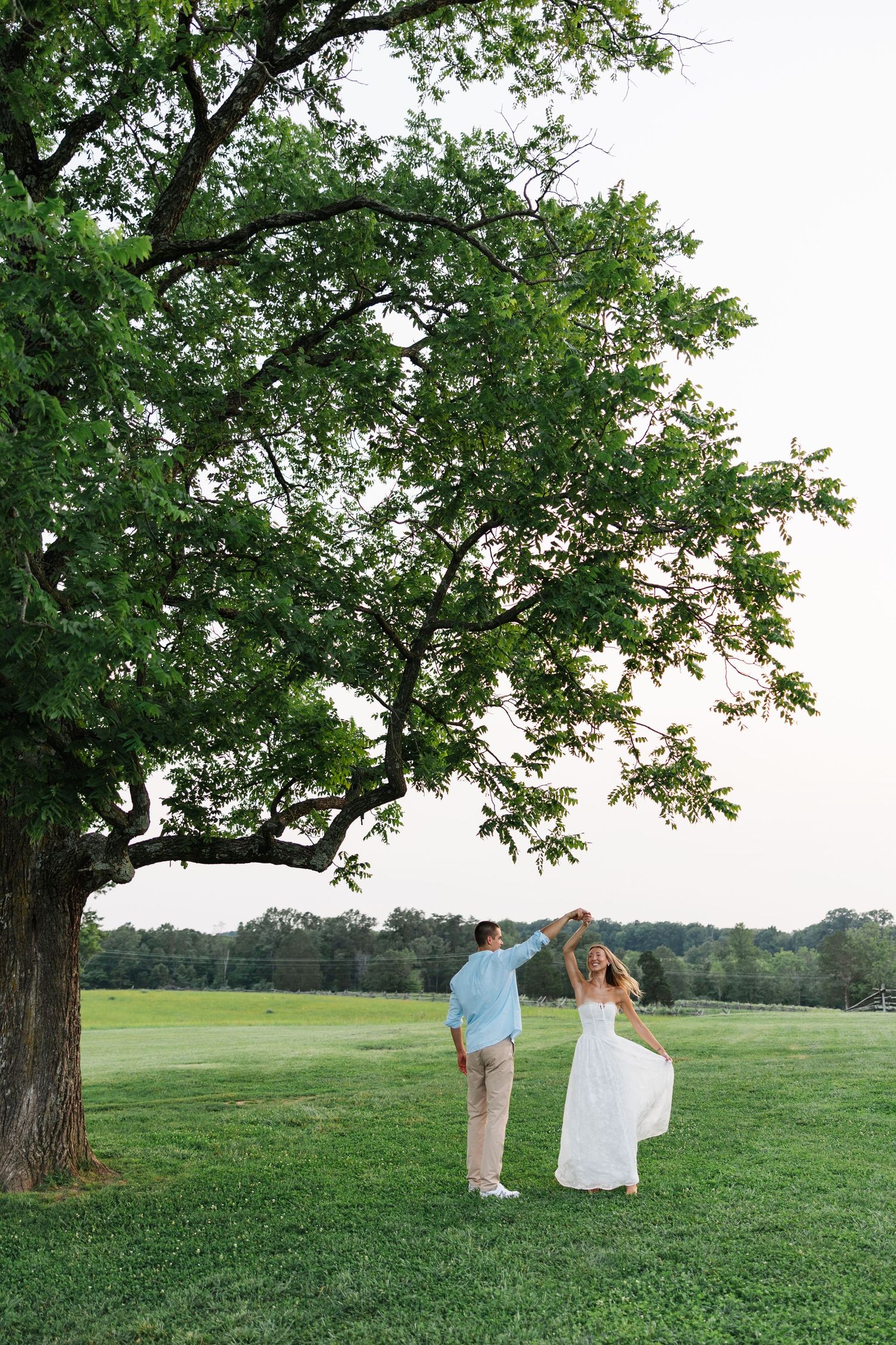 Couple dancing under a large oak tree in a green field, woman in white dress, man in light blue shirt and khakis.