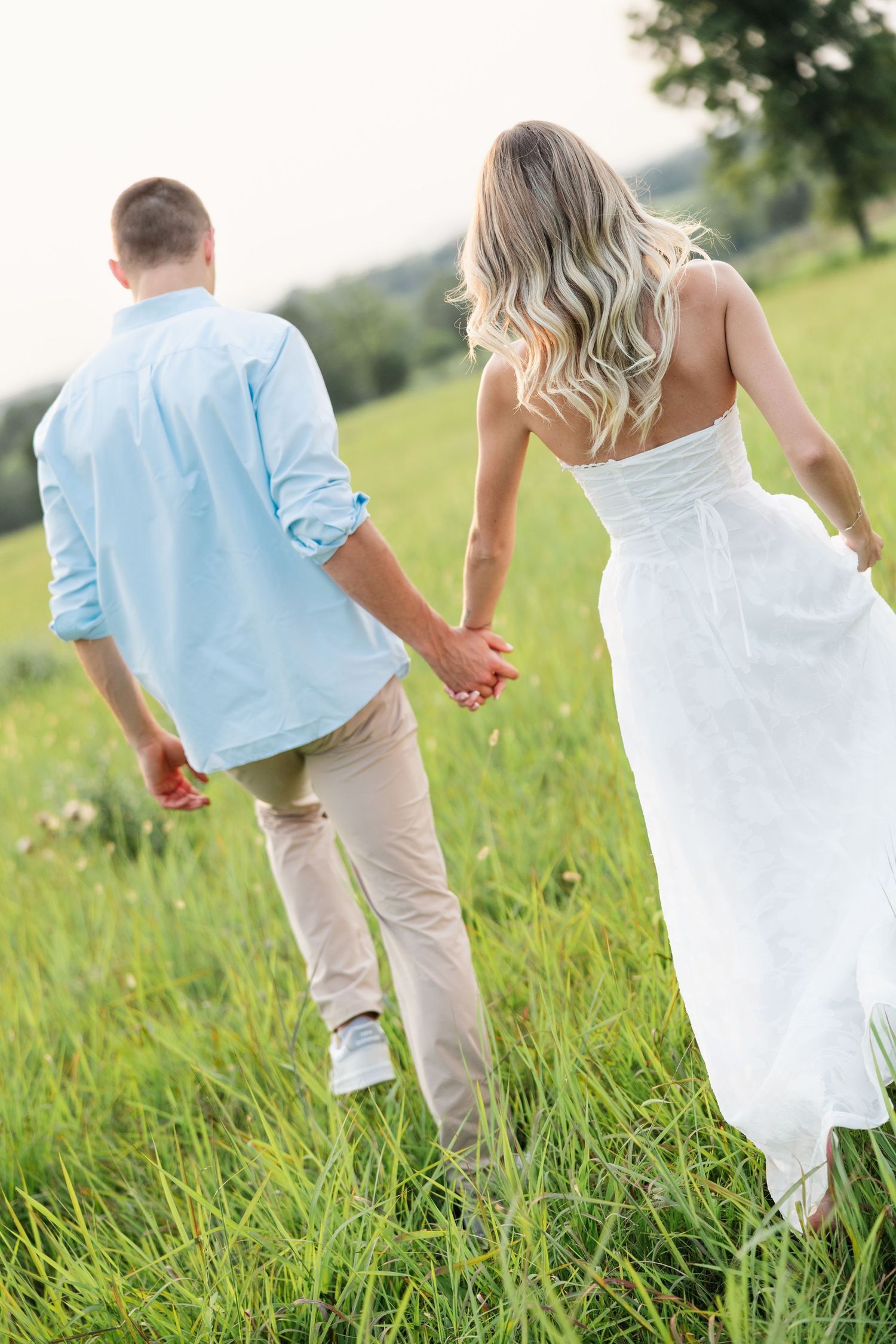 Couple holding hands walking through a lush green field, woman in white dress and man in blue shirt.
