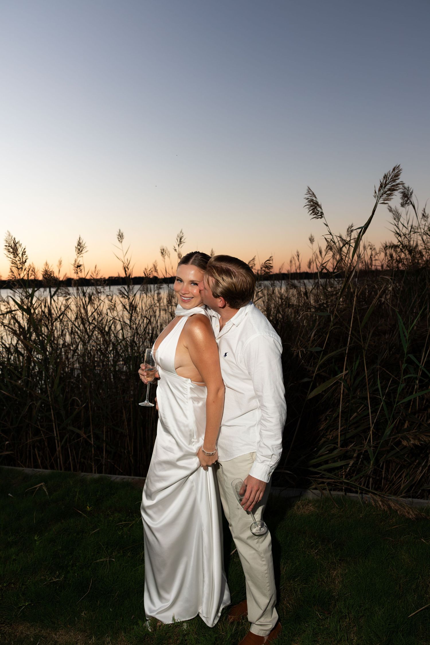 Couple in white attire embracing at sunset near reeds and water, woman holding champagne glass.