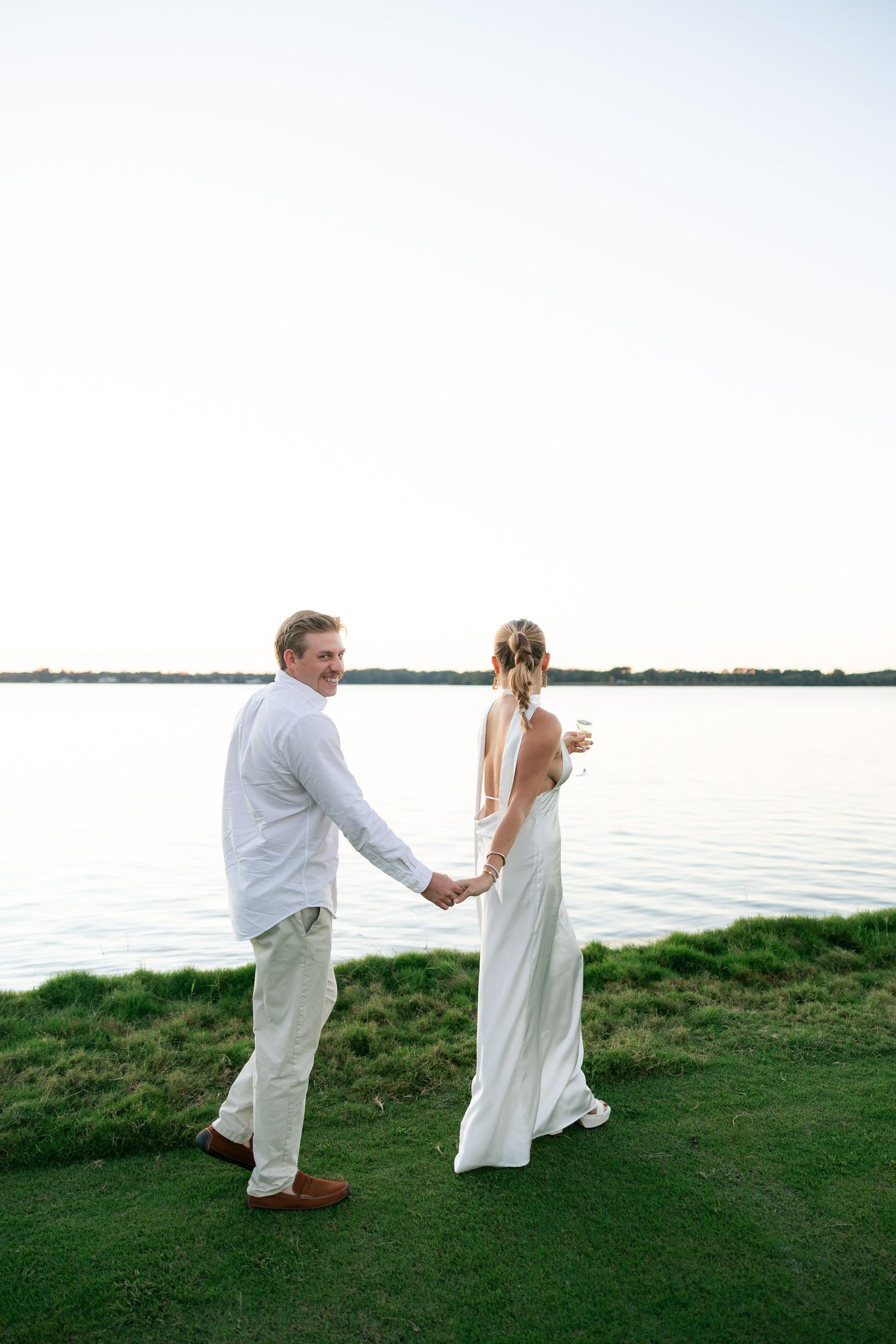 A smiling groom in white leads his bride by the hand toward a serene waterfront at sunset.