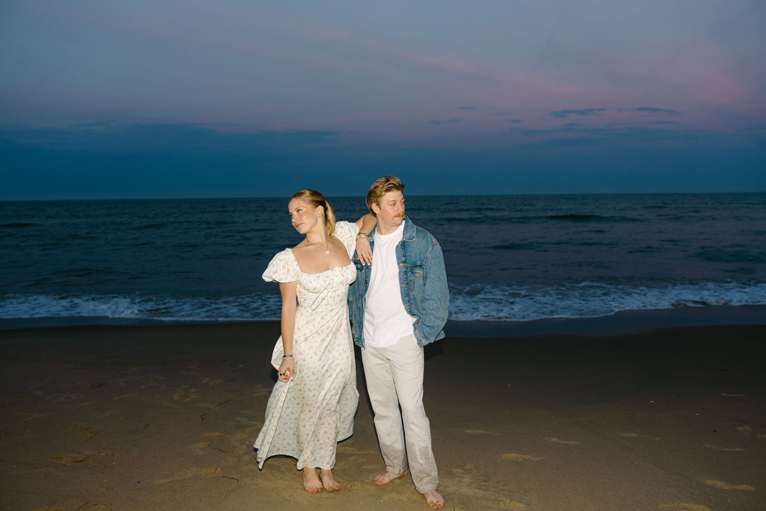Couple walks barefoot on beach together under vibrant pink and blue evening sky.
