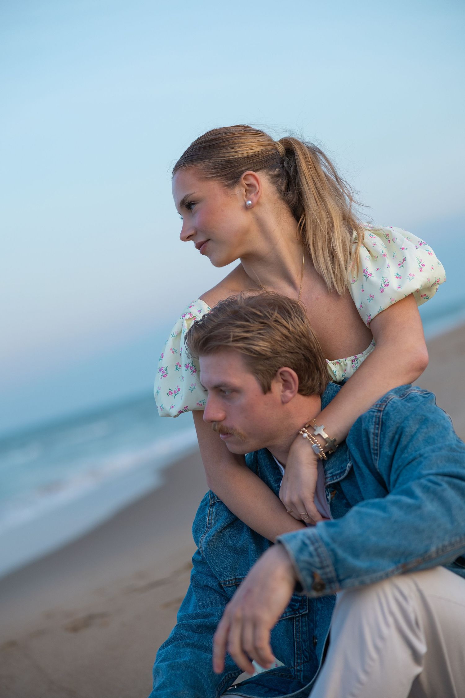 Couple sitting on beach at twilight, woman resting chin on mans shoulder gazing at ocean.