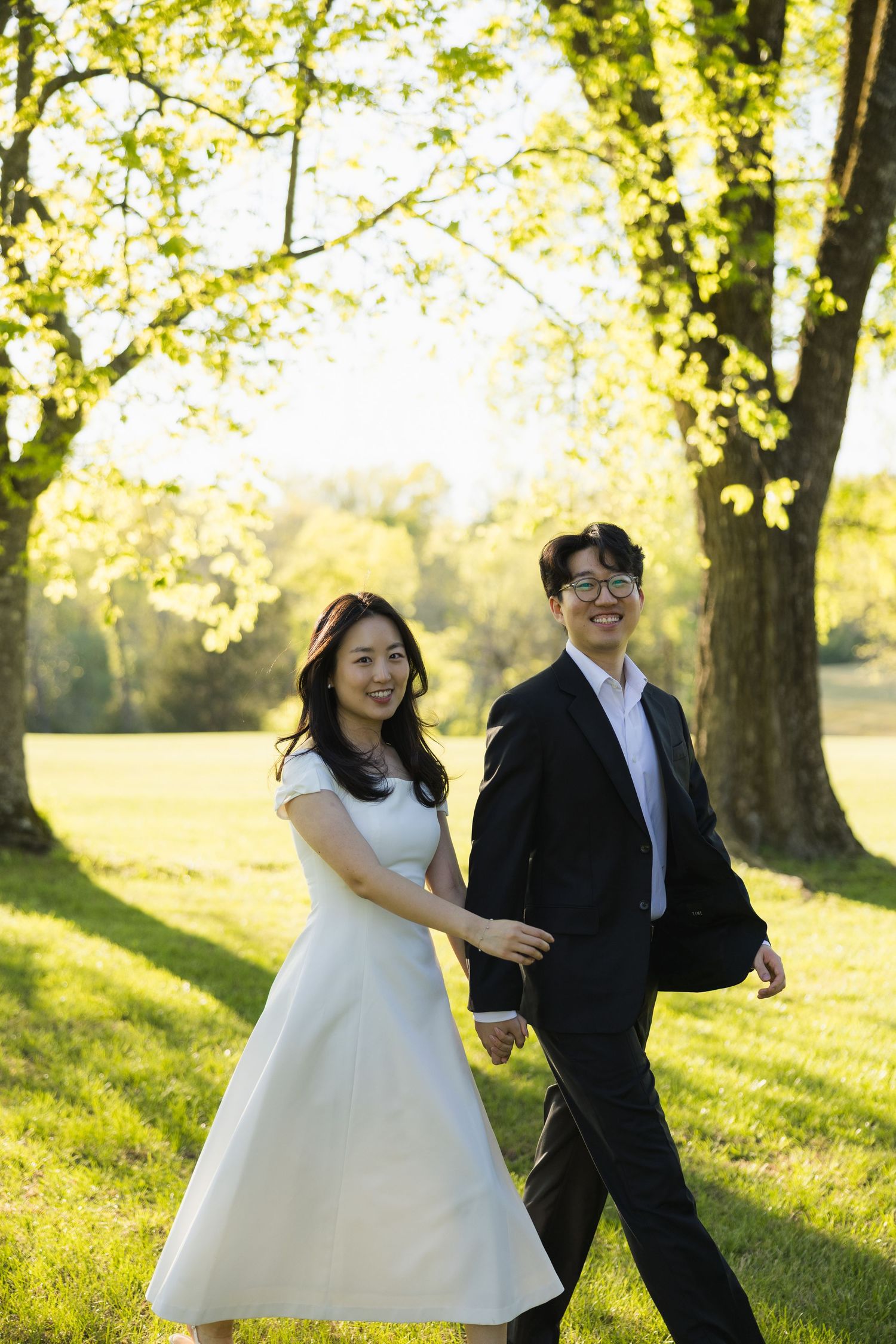 Smiling Asian couple holding hands walking in sunlit green park.