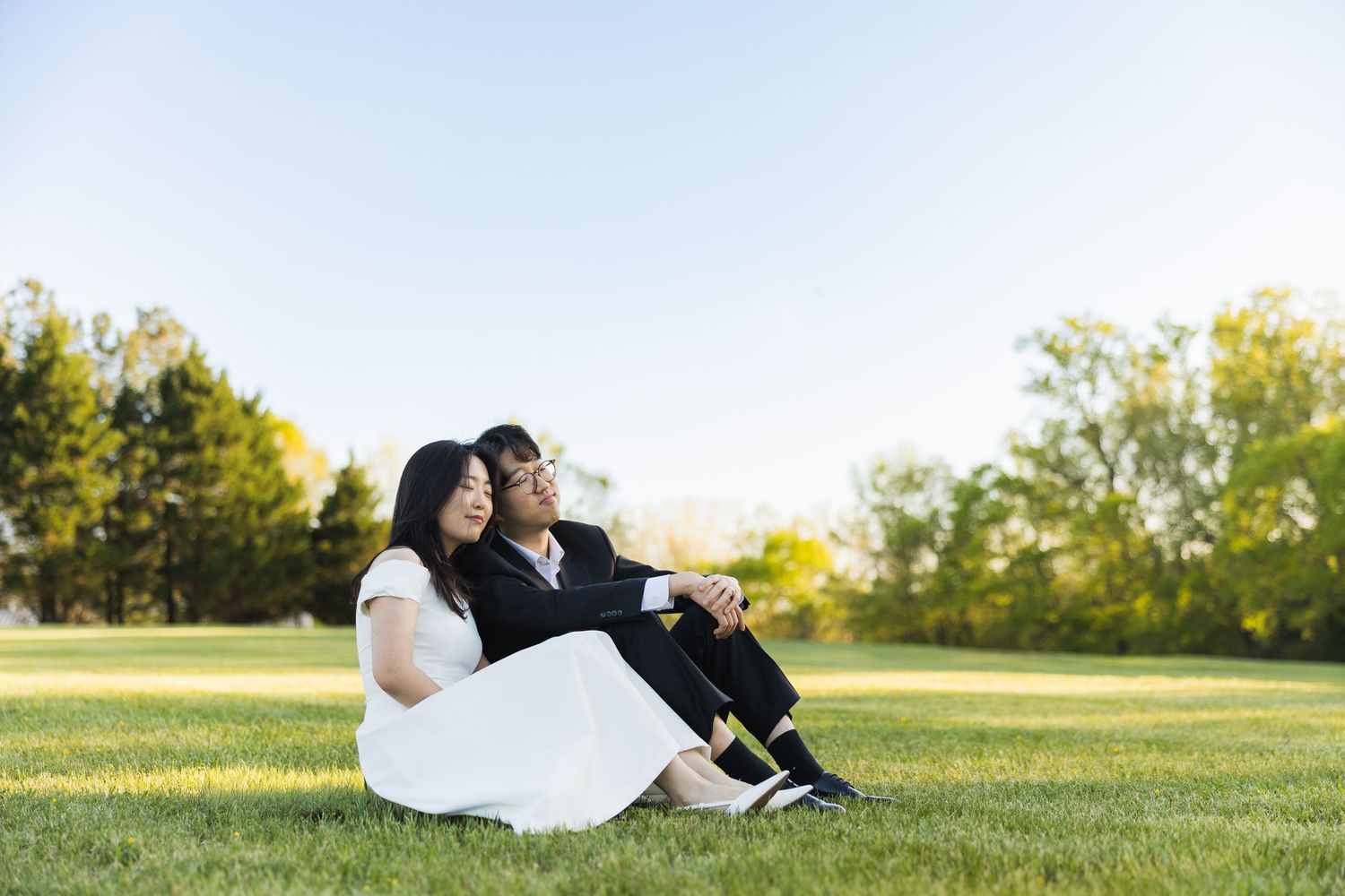 Couple in wedding attire sitting together on green grass beneath a clear blue sky at sunset.