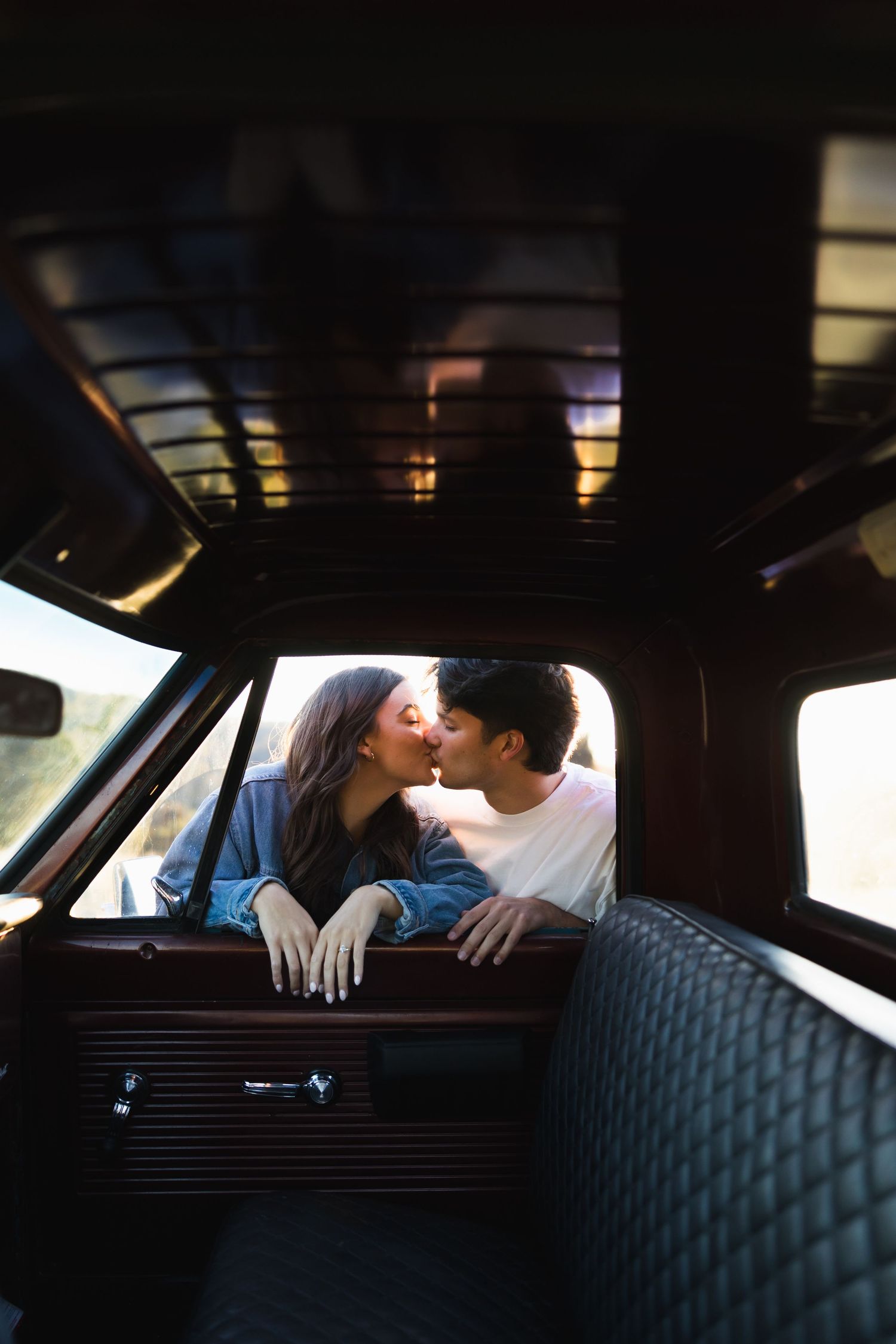 Couple sharing a tender kiss leaning over the back seat of a vintage classic car interior at sunset.
