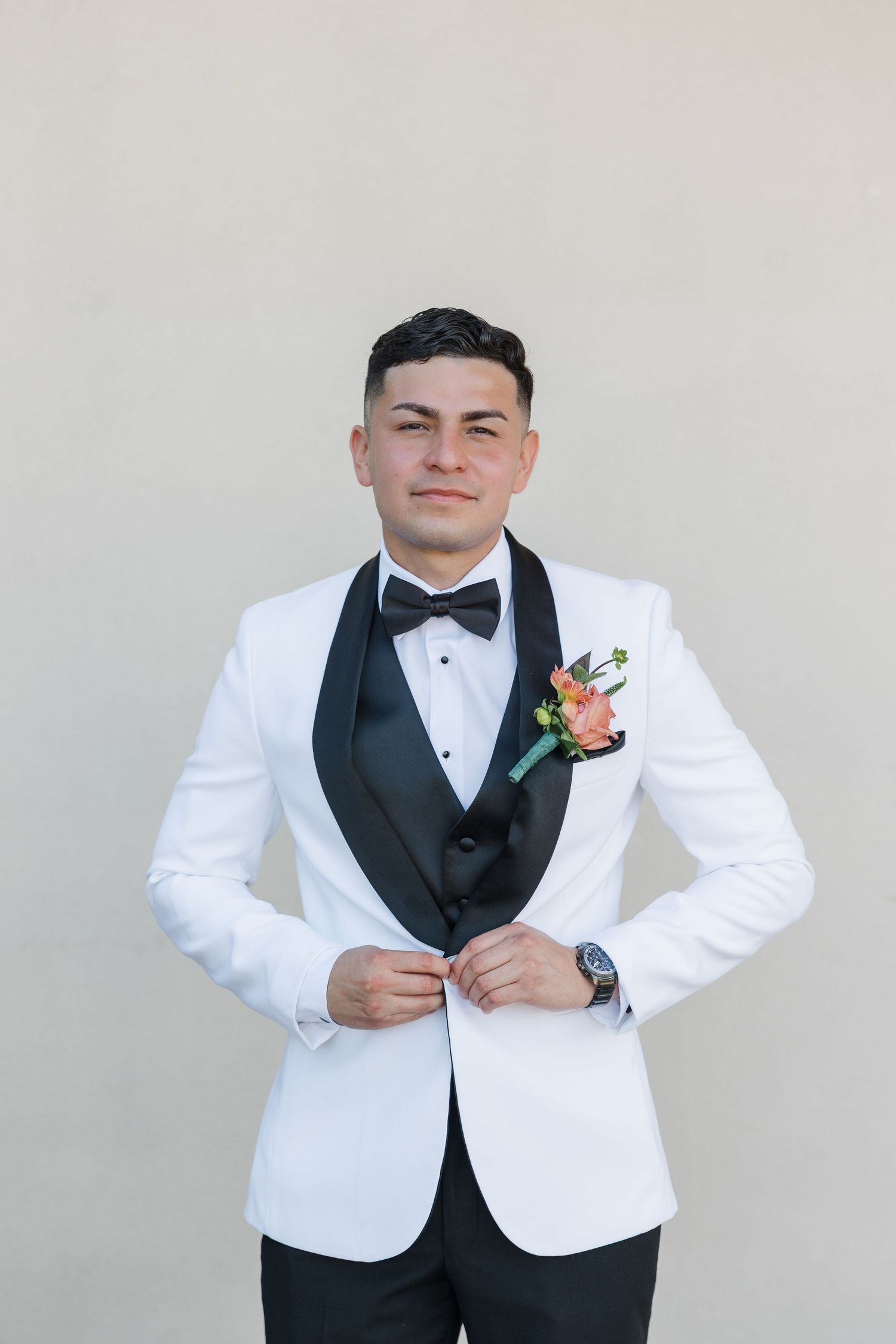 Young groom in white tuxedo with black bow tie and floral boutonniere posing confidently for wedding portrait.
