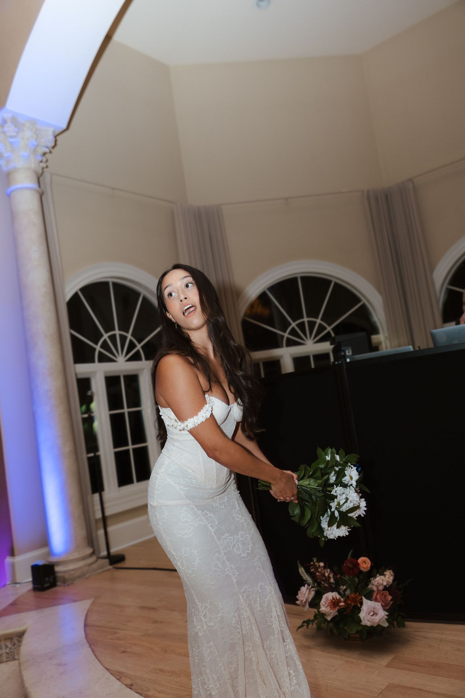Bride in white lace gown preparing to toss bouquet at elegant wedding reception venue.