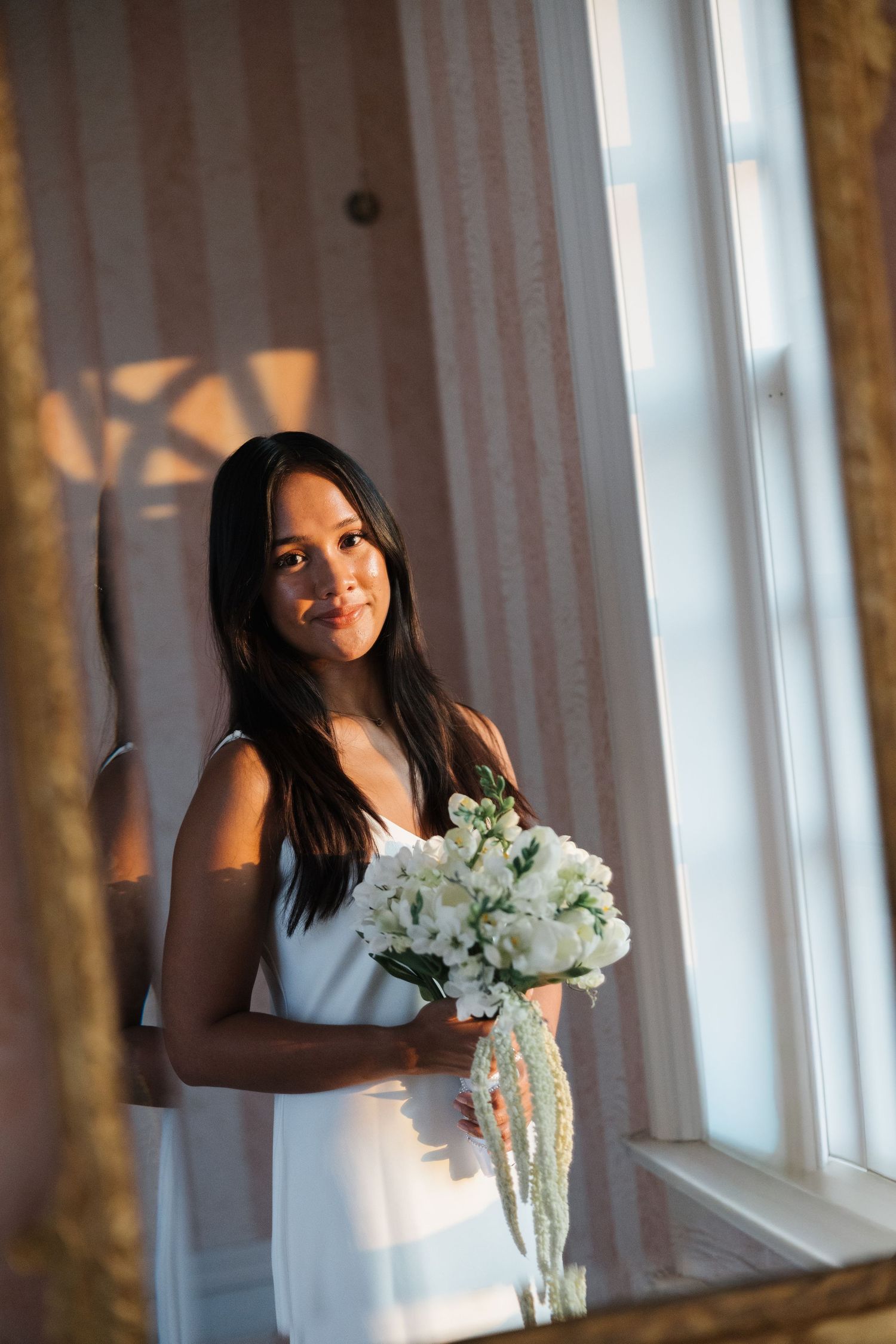 Young woman in white dress holding a bouquet of white flowers, standing by a sunlit window.